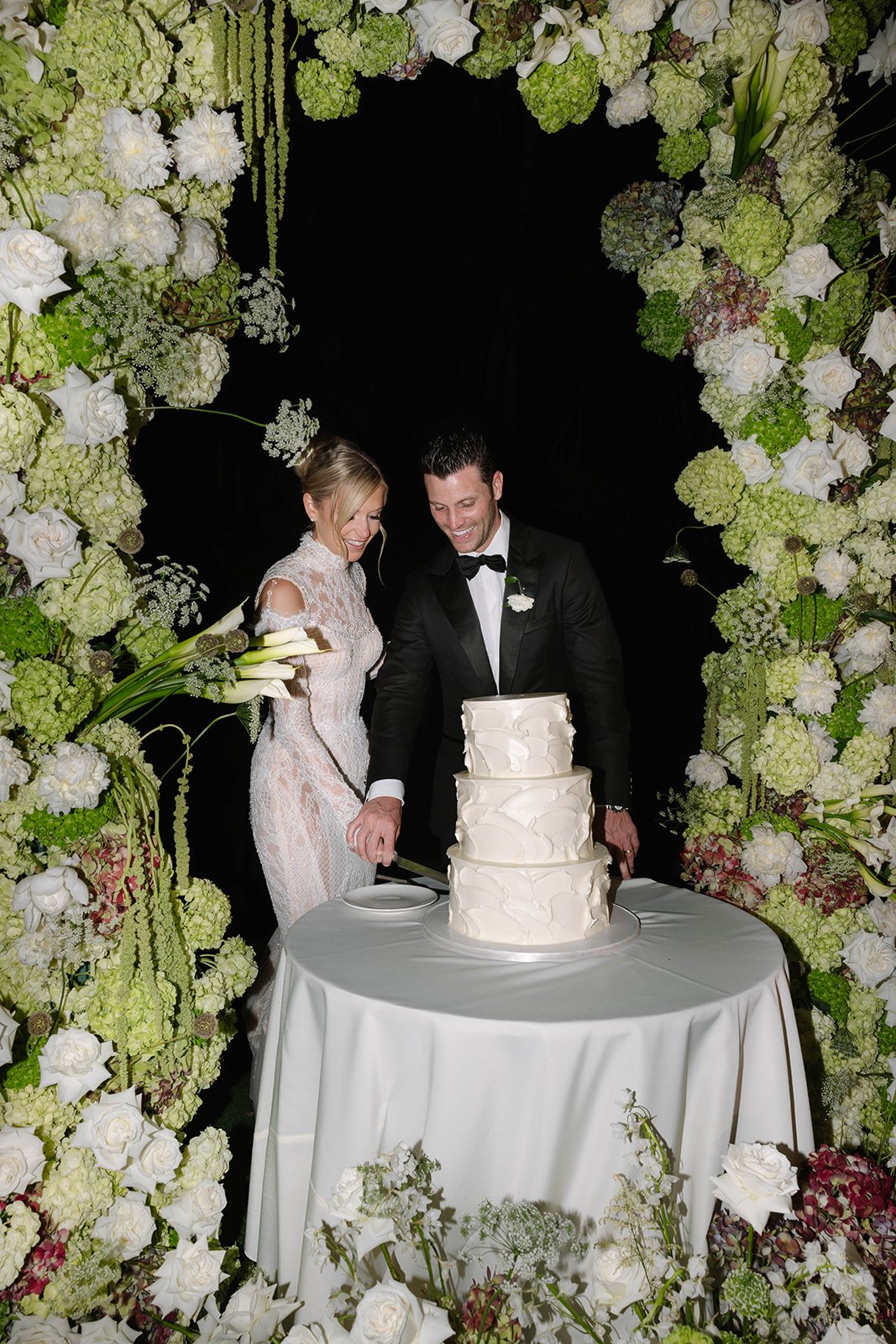 A newly married couple cutting a white wedding cake surrounded by a floral arch of white and green flowers.