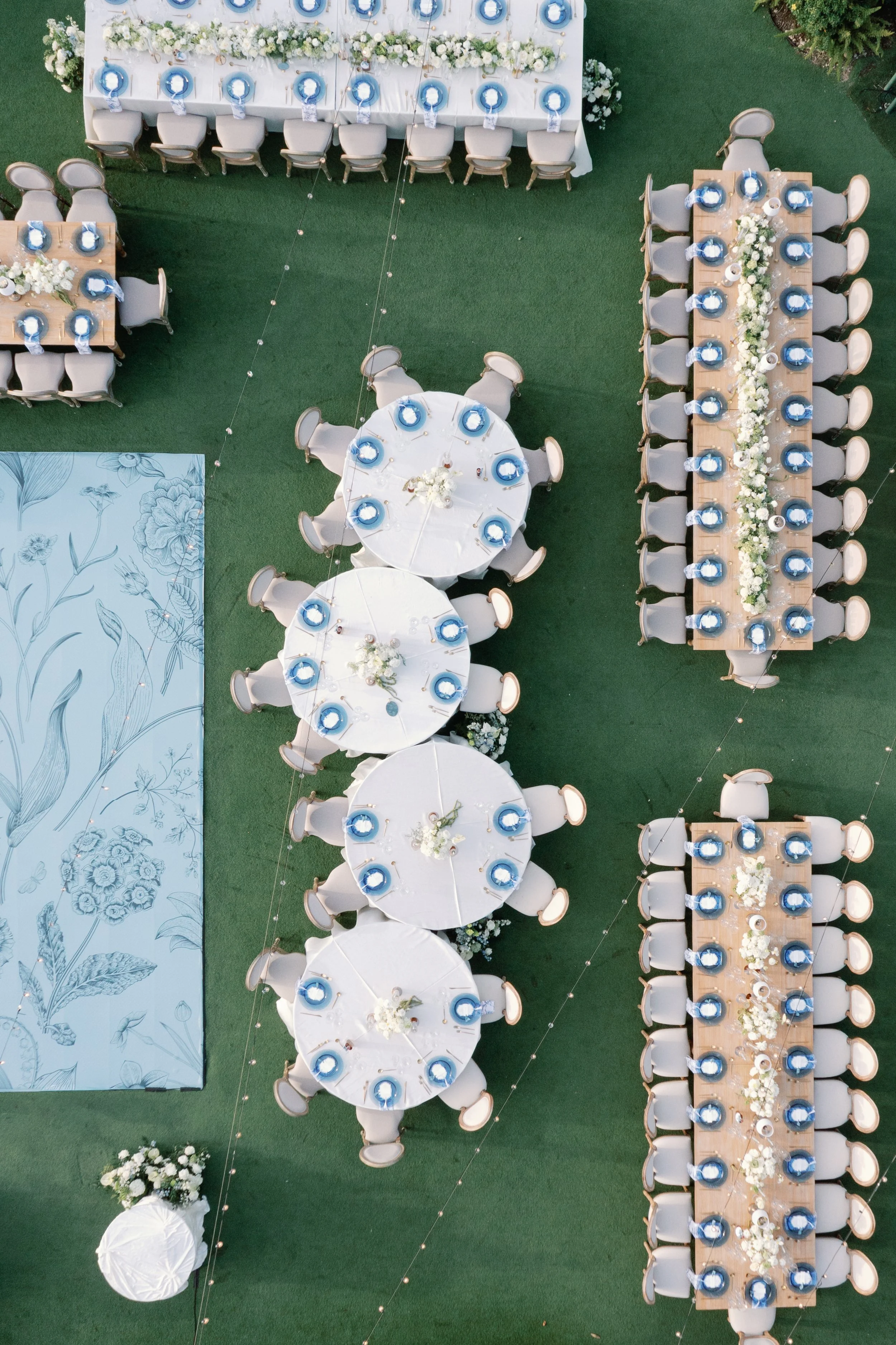Arrangement of decorated tables and chairs for a wedding or event, with white and blue color theme, floral centerpieces, and outdoor setting on green ground.