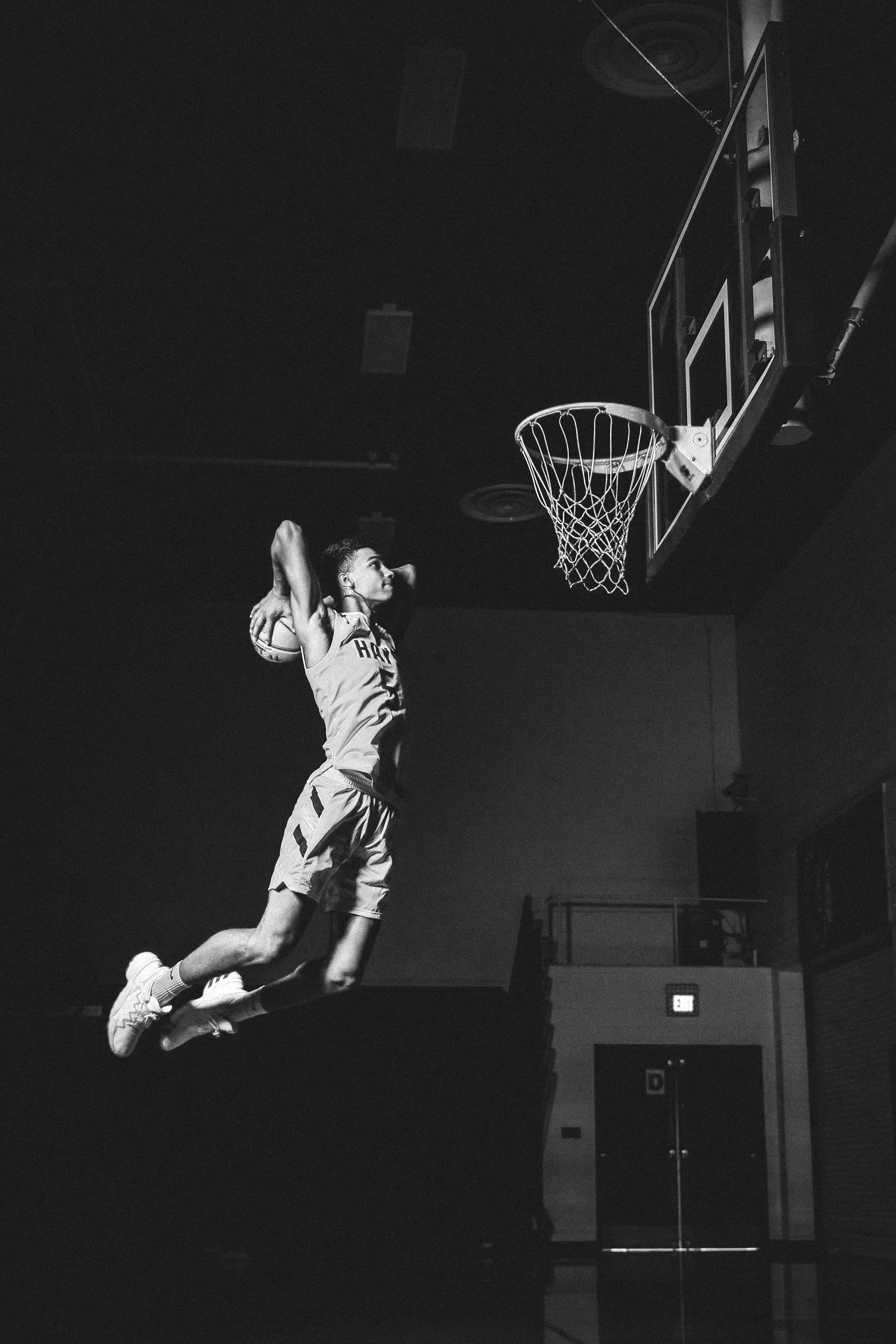 Un joueur de basketball en plein saut vers le panier dans une salle de sport, vue en noir et blanc.