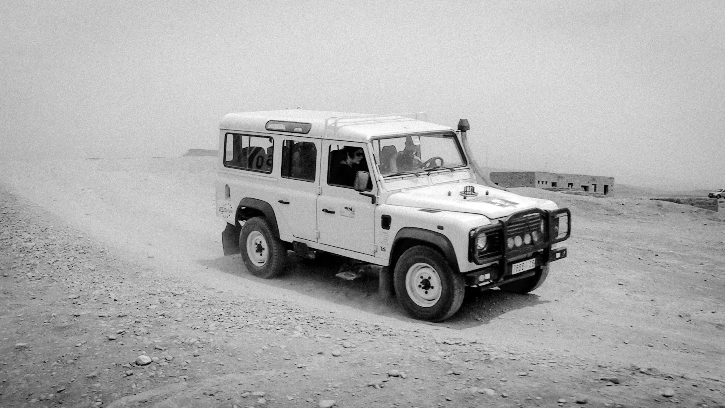 Voiture tout-terrain blanche roulant sur une route poussiéreuse dans un désert, avec un bâtiment en arrière-plan.