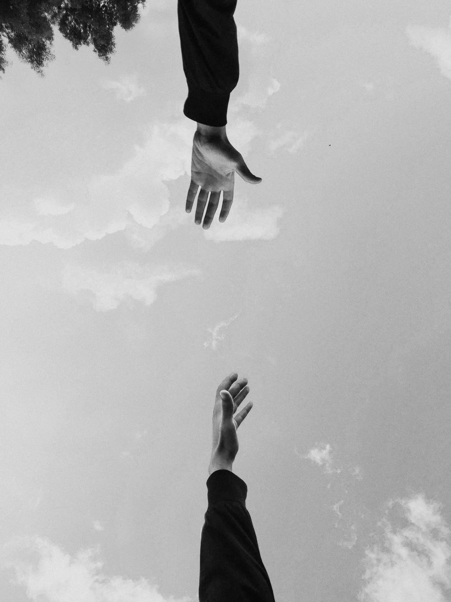 Deux mains s'étendant vers le ciel, dont une semble donner un coup de main à l'autre, contre un fond de ciel nuageux, en noir et blanc.