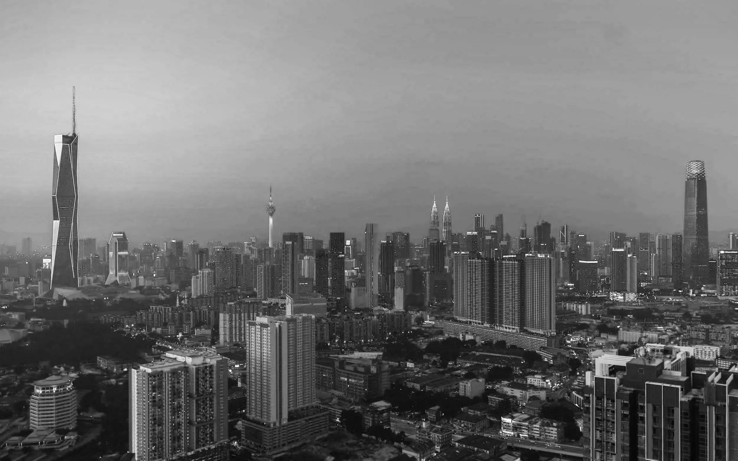Une vue en monochrome d'une ville moderne avec de nombreux gratte-ciel, dont le plus remarquable est la tour avec des formes géométriques audacieuses à gauche, et deux célèbres tours jumelles dans le centre, probablement Kuala Lumpur, avec d'autres gratte-ciel en arrière-plan.
