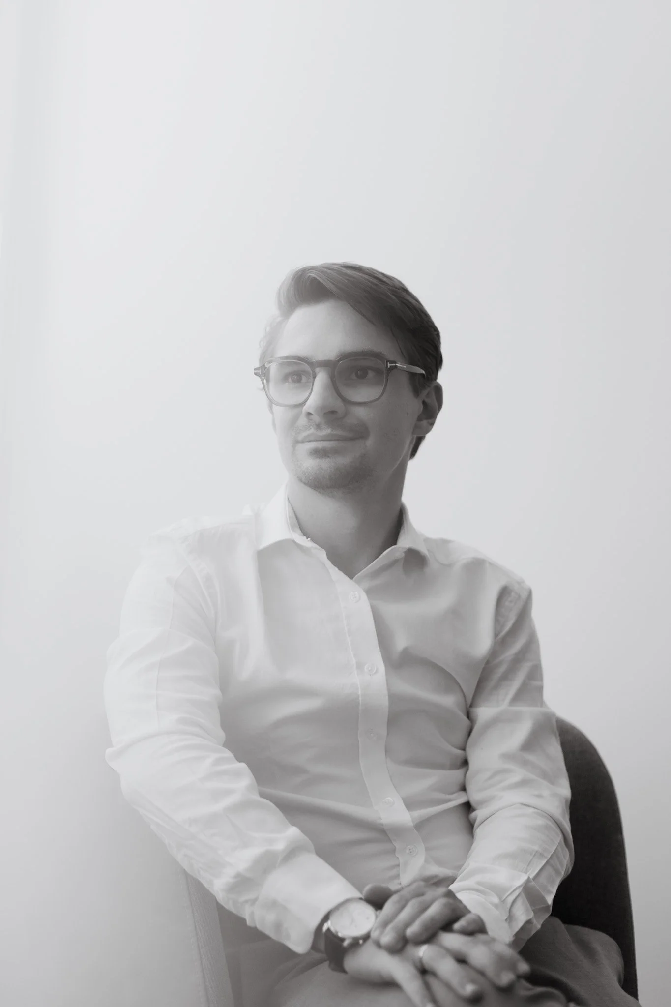Correntin Boix-Vives. Expert-comptable. Commissaire aux comptes. Homme avec lunettes, chemise blanche, assis dans un fauteuil, regardant vers la gauche, mains croisées, montre au poignet gauche, arrière-plan blanc.