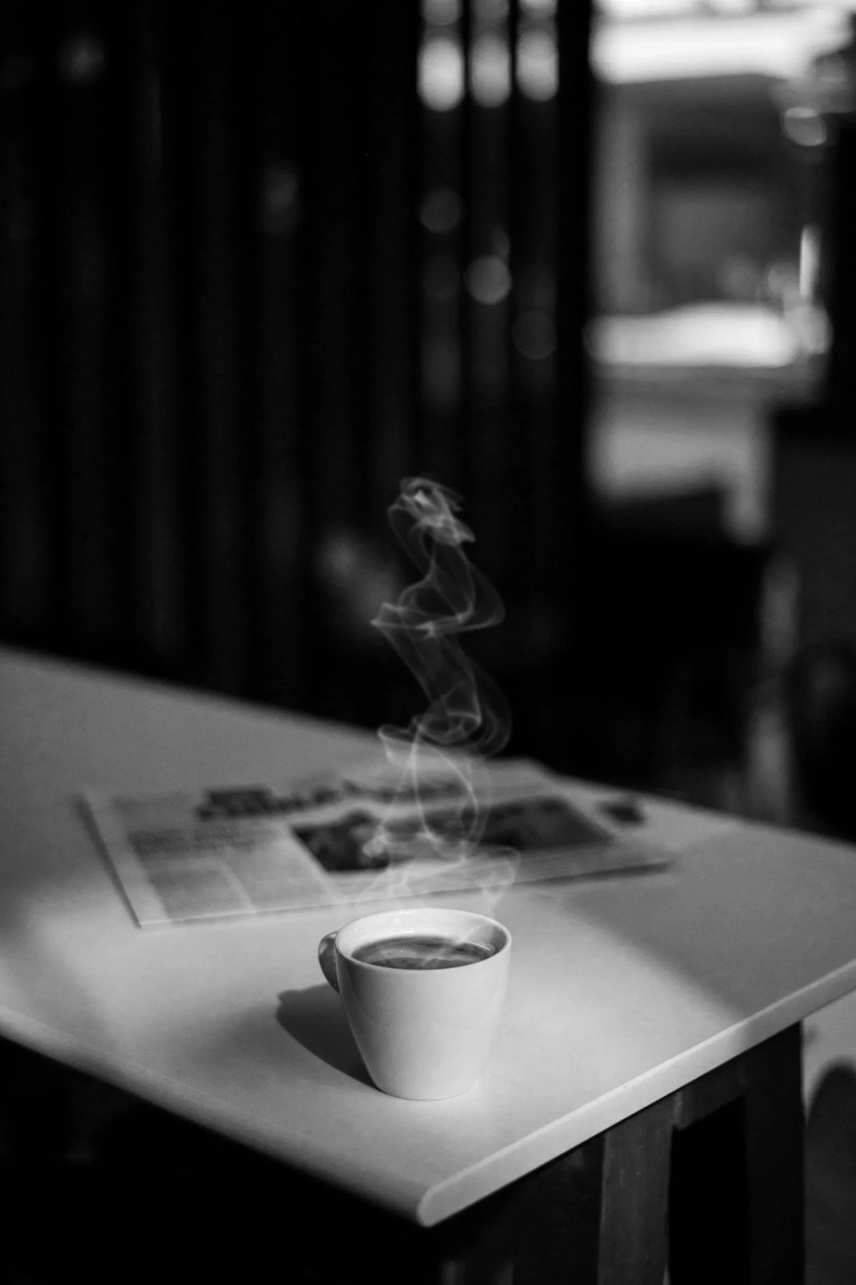 Une tasse de café chaud avec de la vapeur qui s'élève et une nappe blanche. En arrière-plan, une table avec des documents ou un magazine, dans un environnement intérieur avec une fenêtre en arrière-plan laissant entrer la lumière.