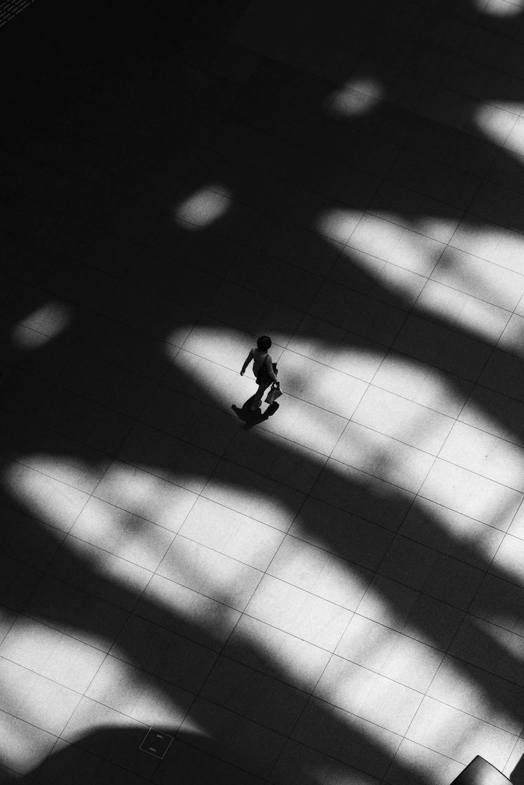 Une personne marche dans un espace intérieur avec des ombres sur le sol, photographié en noir et blanc.