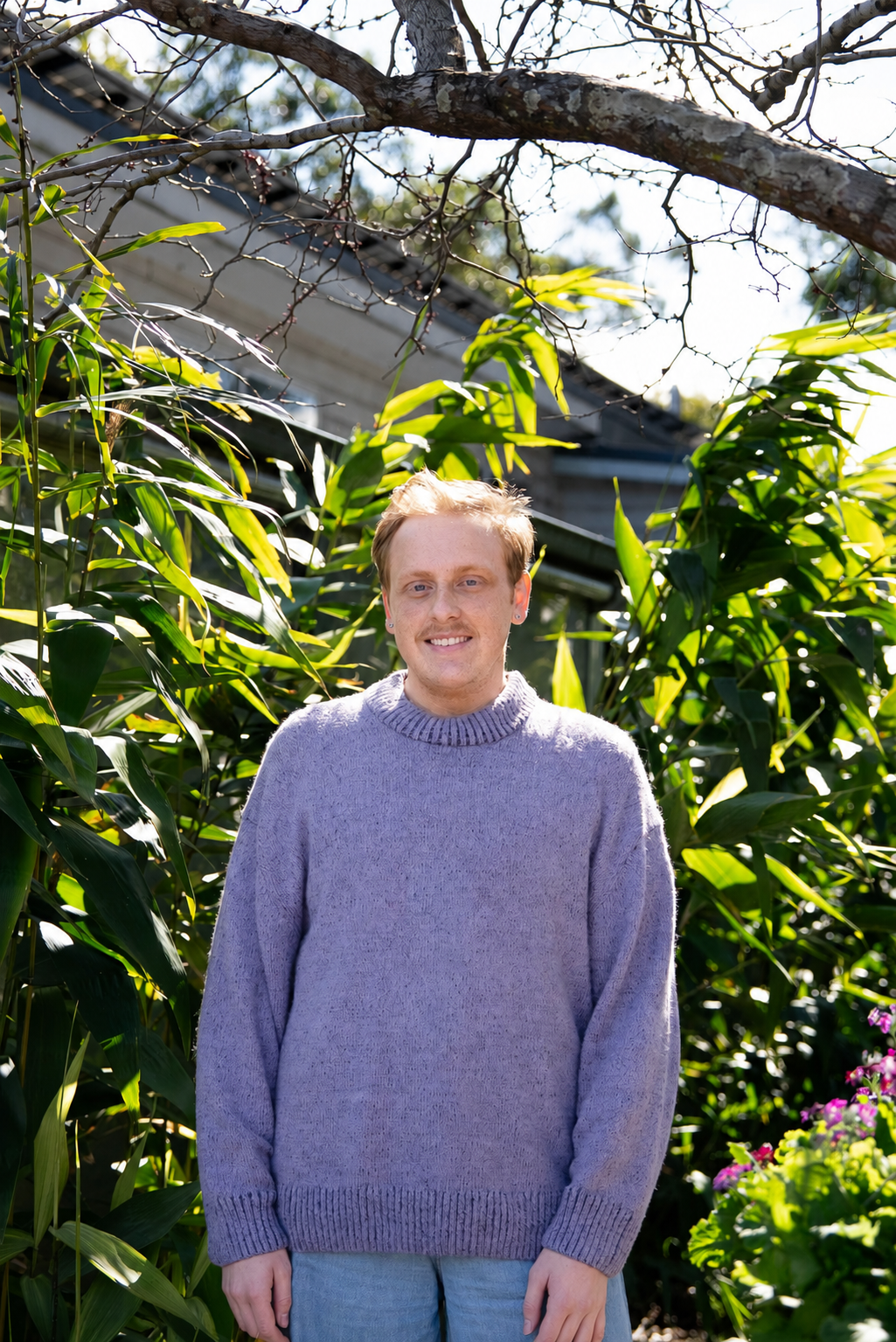 A young man with blond hair and a light purple sweater standing in a lush garden with tall green plants and trees.