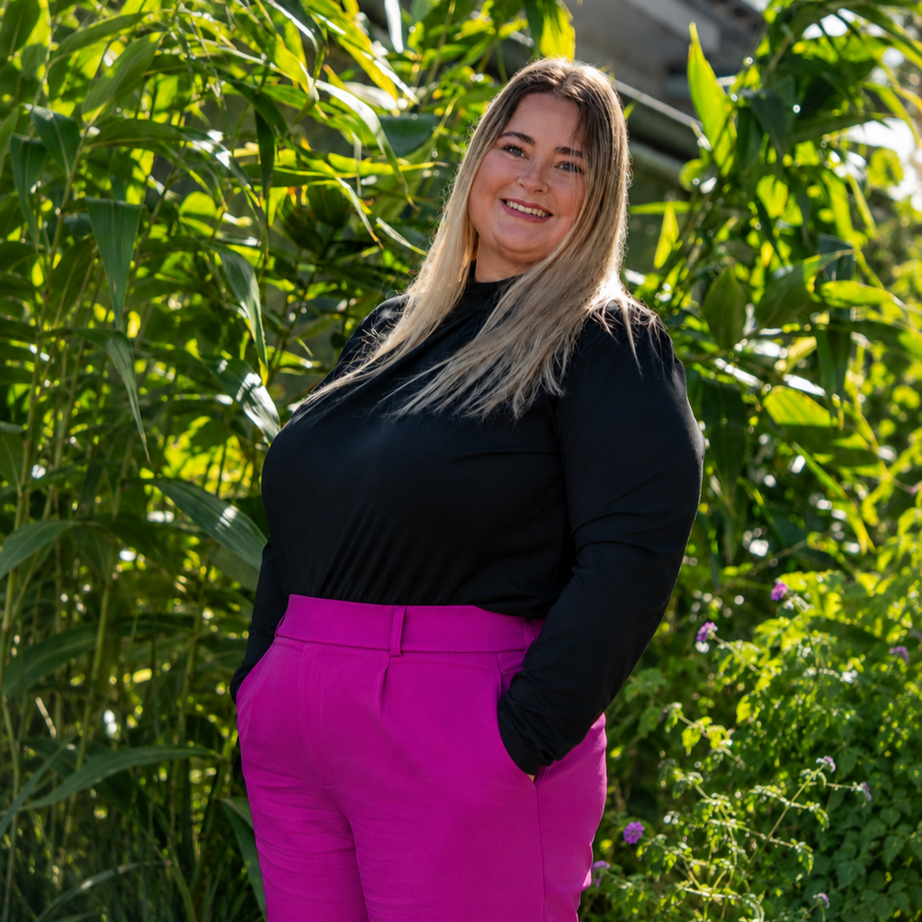 A woman with long blonde hair wearing a black top and pink pants standing outdoors among green plants and smiling at the camera.