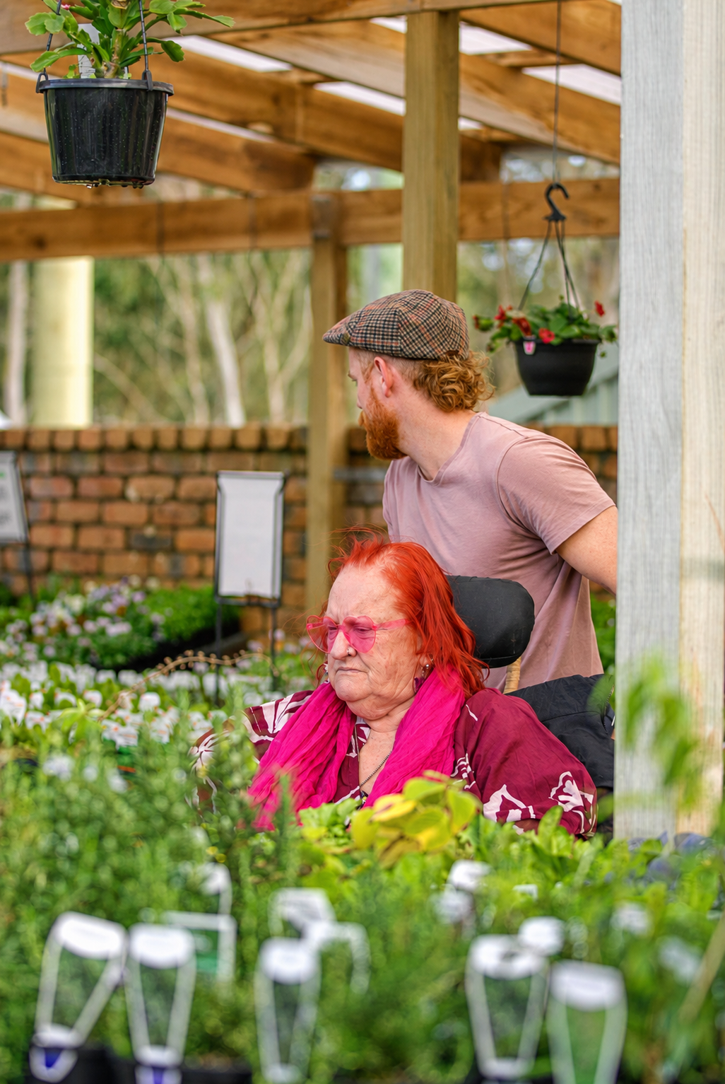 A woman with bright red hair and pink glasses sitting in a wheelchair, surrounded by plants in a garden center. A man with red hair and beard, wearing a cap and pink t-shirt, stands behind her looking to the side. There are hanging flower pots and a brick wall in the background.