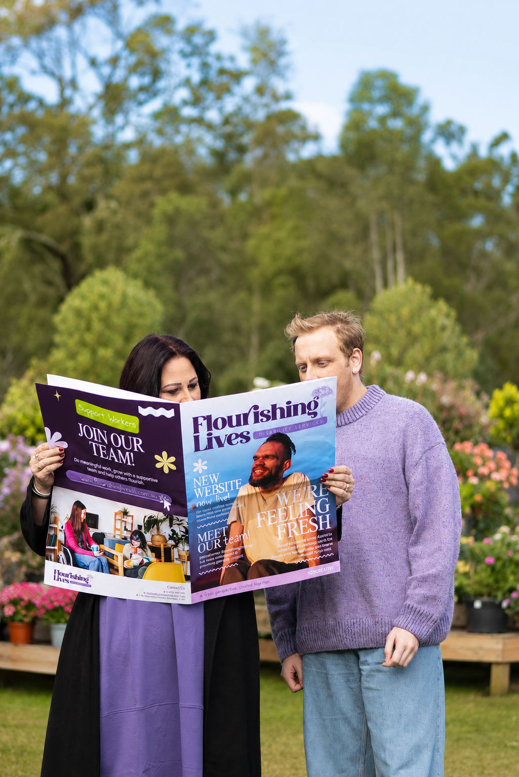 Two people standing outdoors, reading a colorful magazine with garden plants and trees in the background.