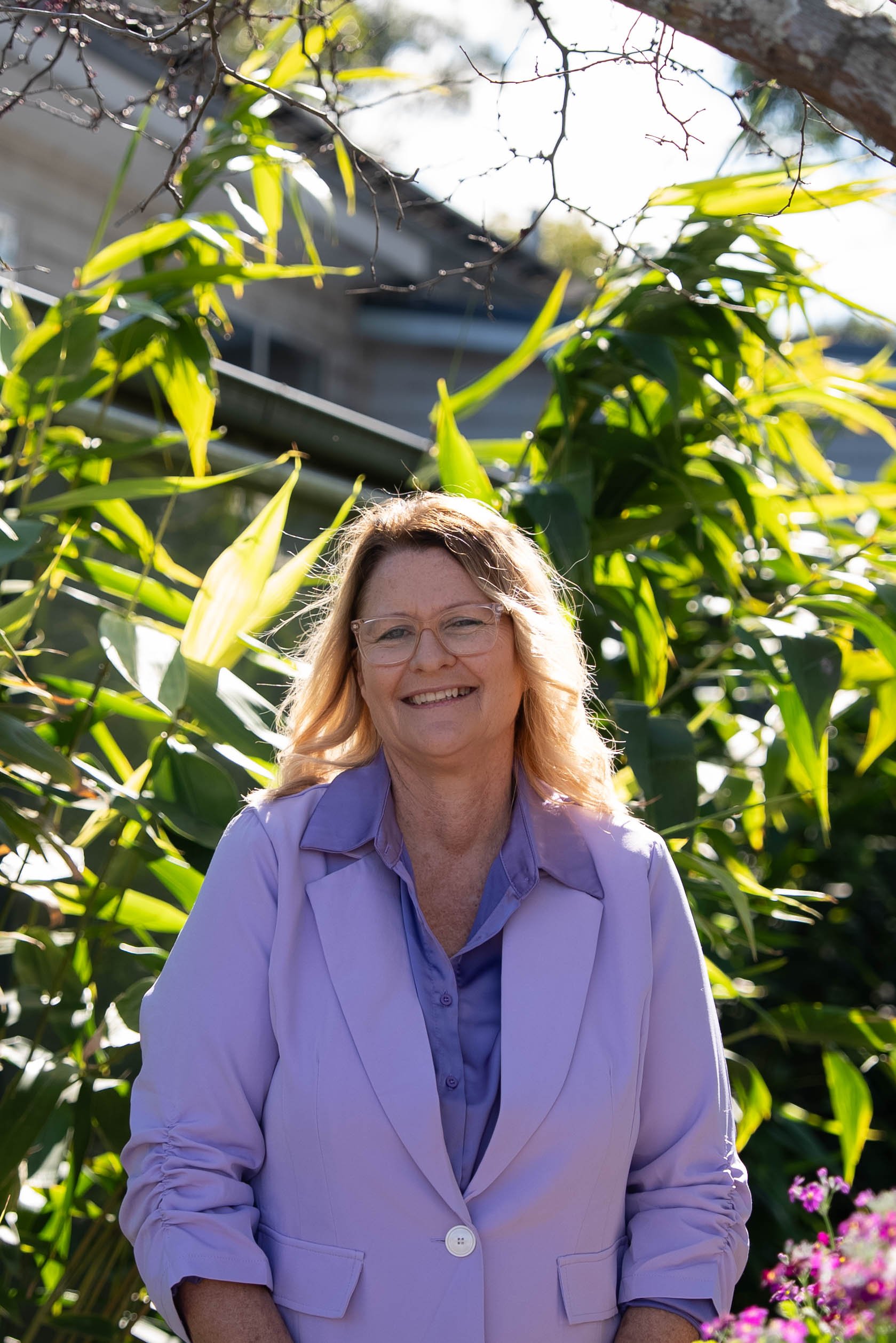 A smiling woman with blonde hair, glasses, wearing a light purple blazer and a purple shirt, standing outdoors in front of green plants and flowers.