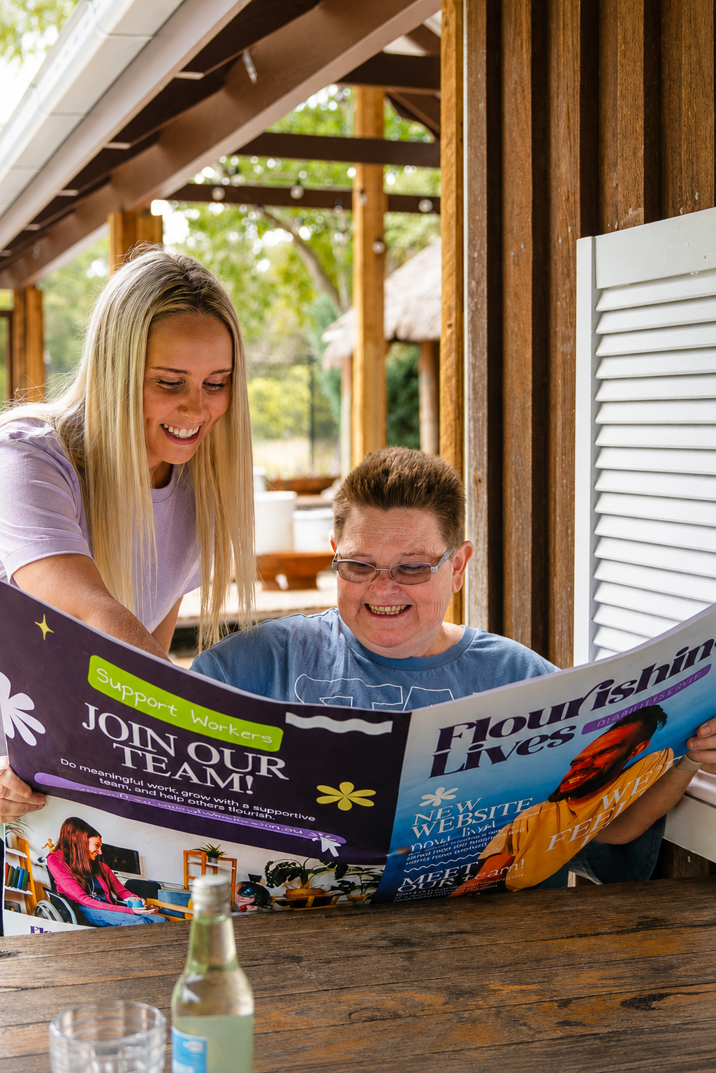A young woman and an older woman are sitting at a wooden table outside. They are both smiling and looking at a colorful brochure. There is a small glass bottle and a glass on the table.