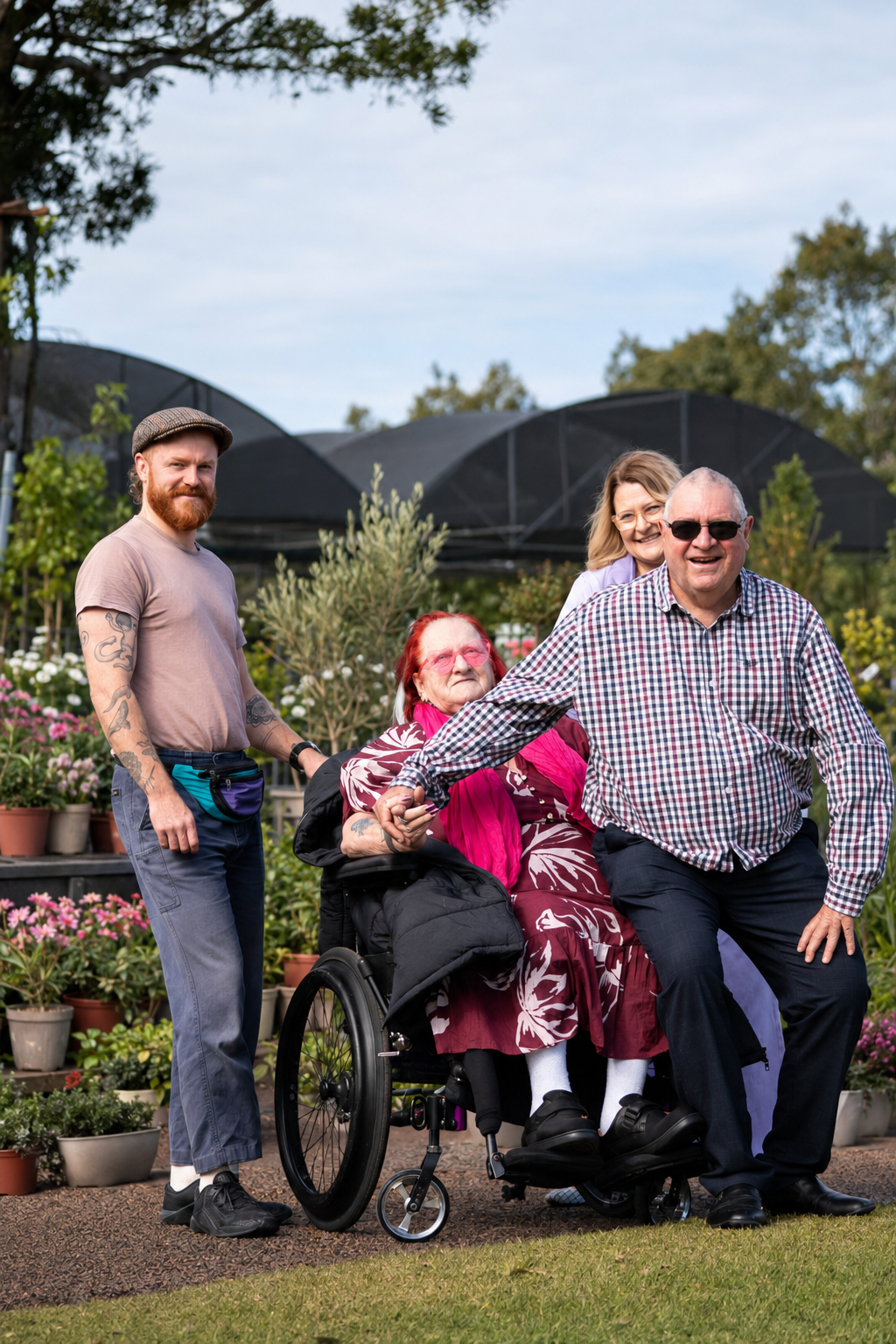 Four people, including an elderly woman in a wheelchair, a man with glasses, a woman, and a young man, are smiling and holding hands outdoors in a garden with potted plants and trees.