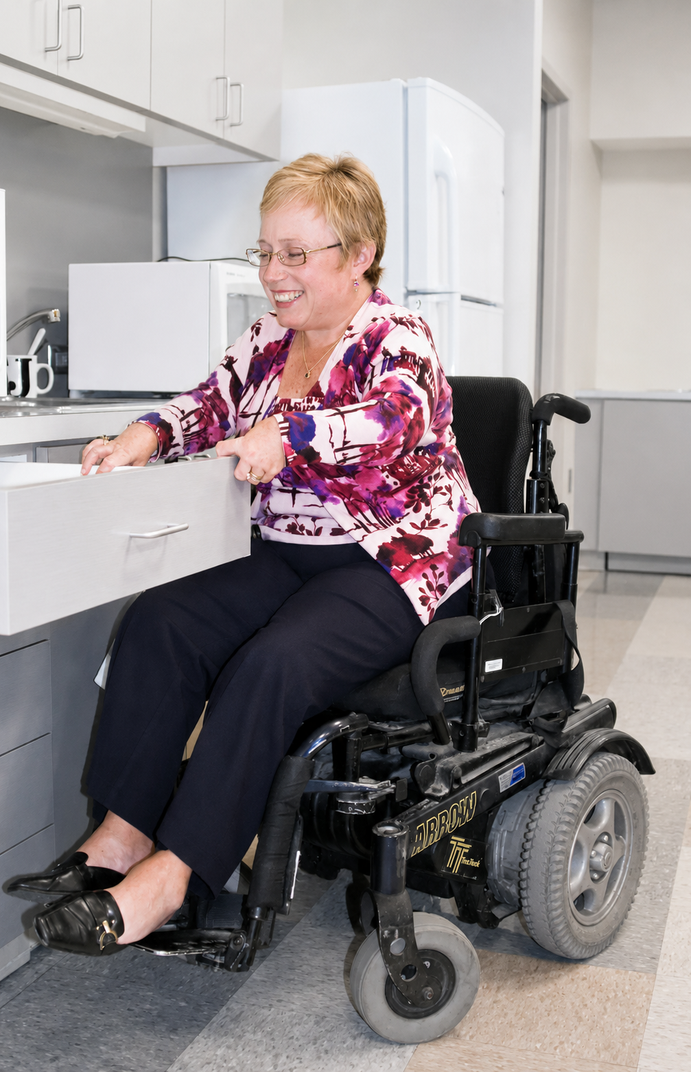 A woman in a floral blouse and black pants sitting in a wheelchair opening a kitchen drawer, smiling.