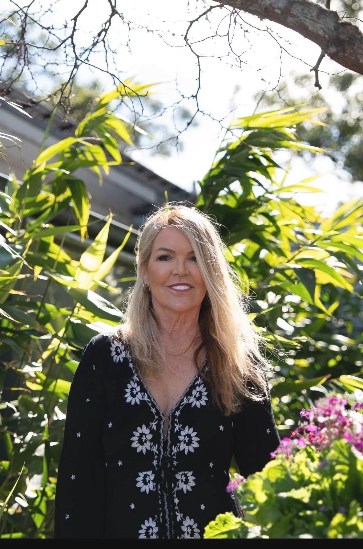 A woman smiling outdoors surrounded by green plants, flowers, and trees, with sunlight illuminating her face.