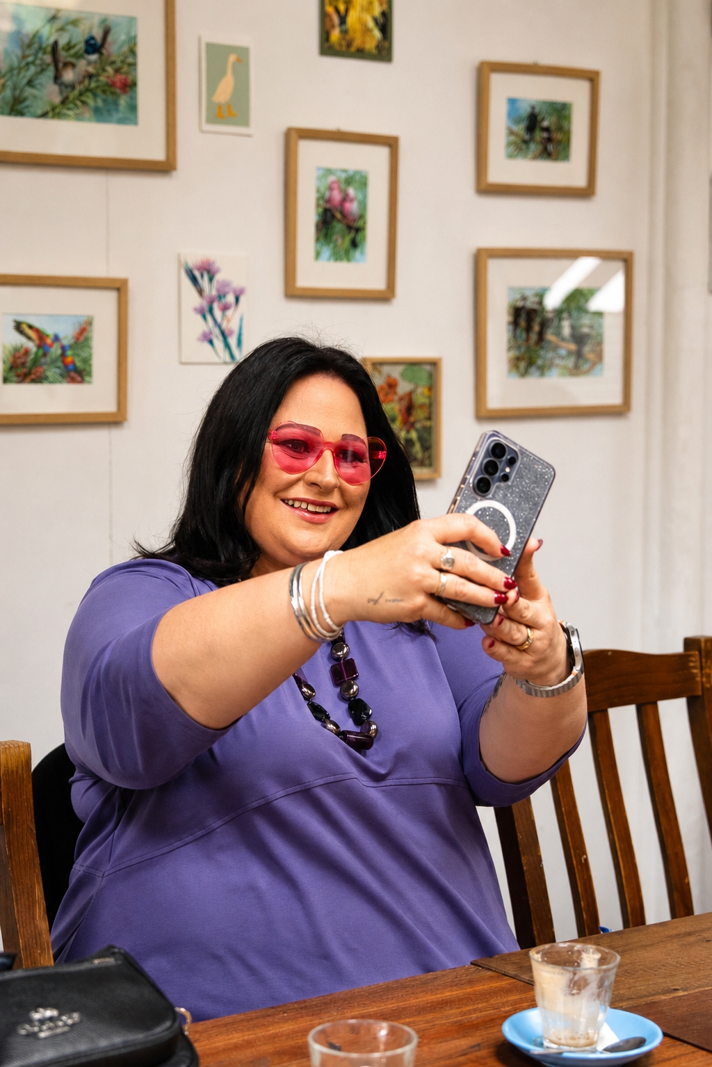 A woman with dark hair and pink sunglasses sitting at a wooden table, smiling and taking a selfie with her phone. The background features a white wall decorated with colorful framed artwork of animals and flowers.
