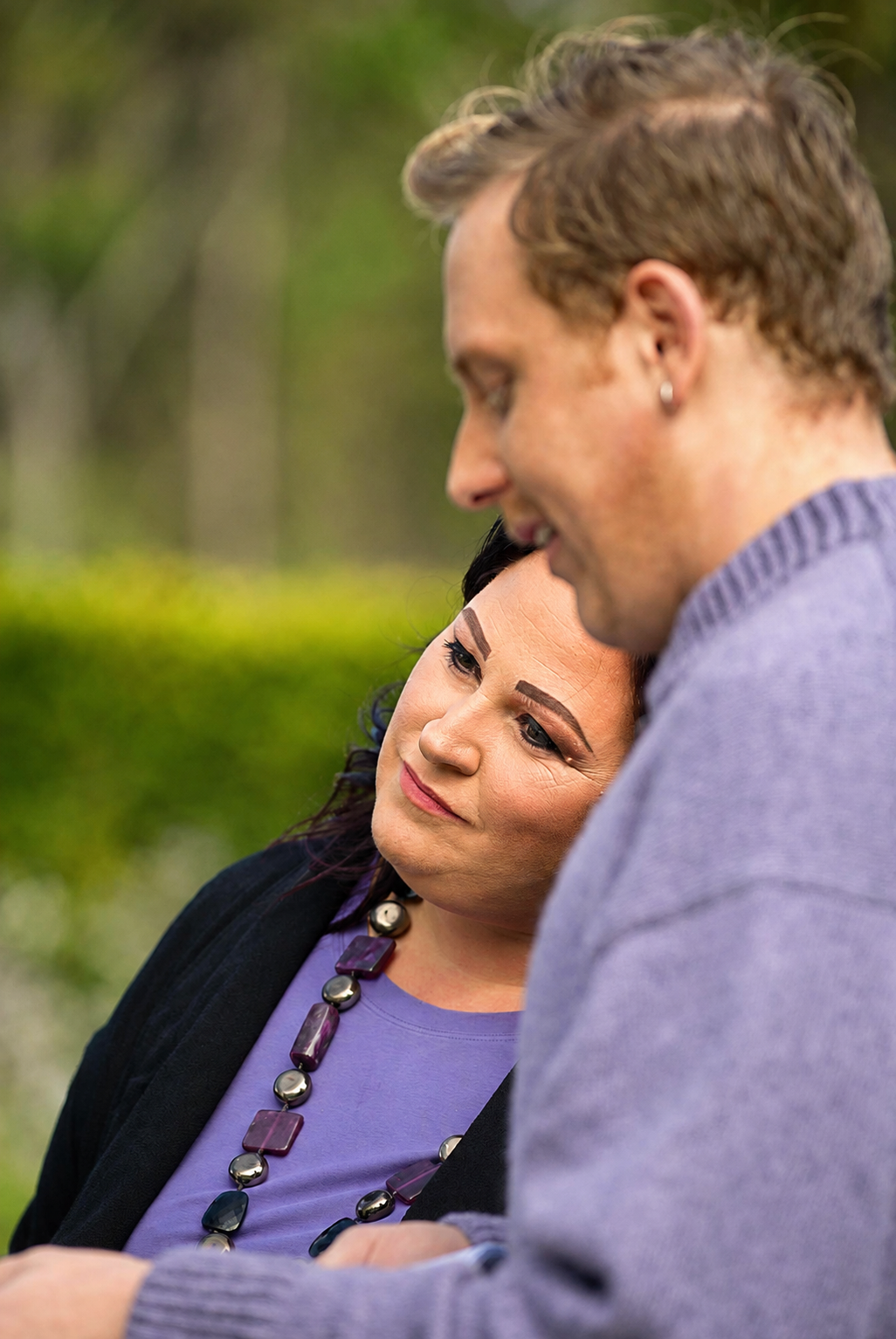 A woman with dark hair, wearing makeup and a purple top with a beaded necklace, is looking at a man with light brown hair, a earring, and a gray shirt, both outdoors with a blurred green background.