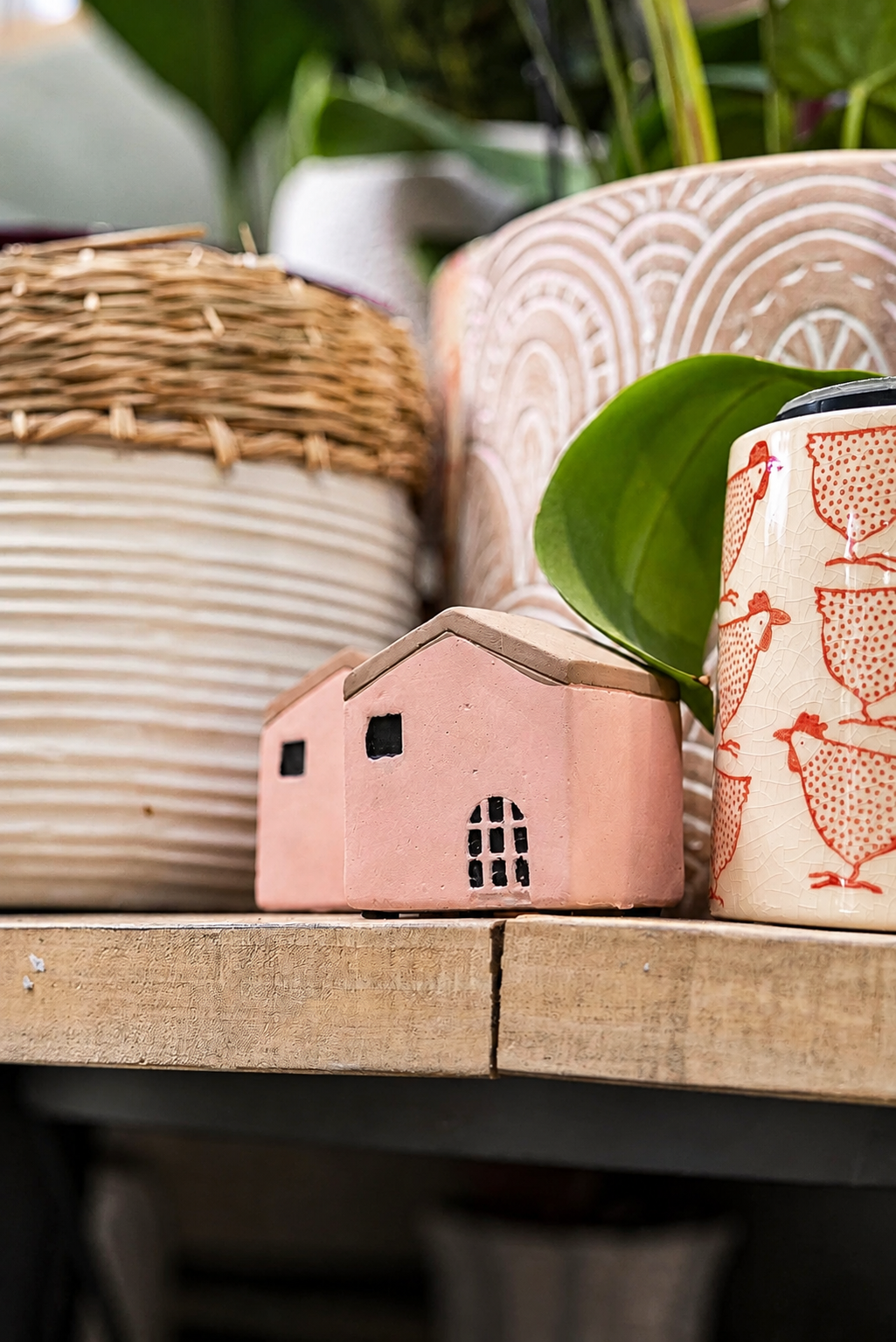 Close-up of small pink house decoration with black windows and door on a wooden surface, surrounded by various patterned pots and a green leaf in the background.