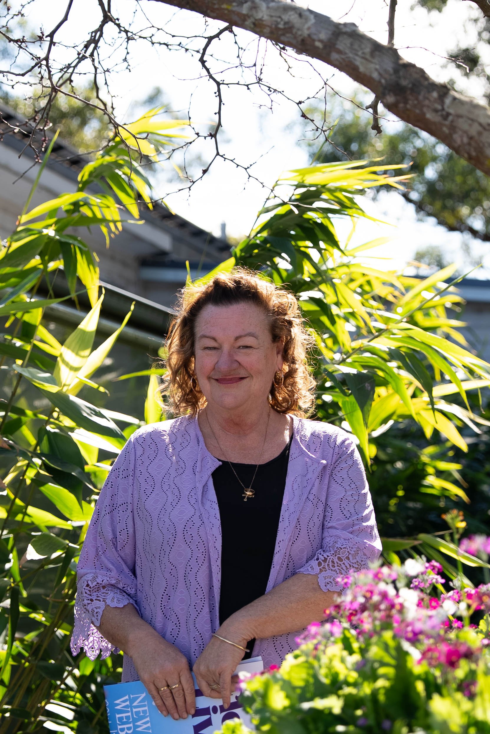 A woman with curly hair wearing a black top and a light purple cardigan standing outdoors in a garden with green plants, flowers, and a tree branch overhead, holding a magazine or pamphlet.