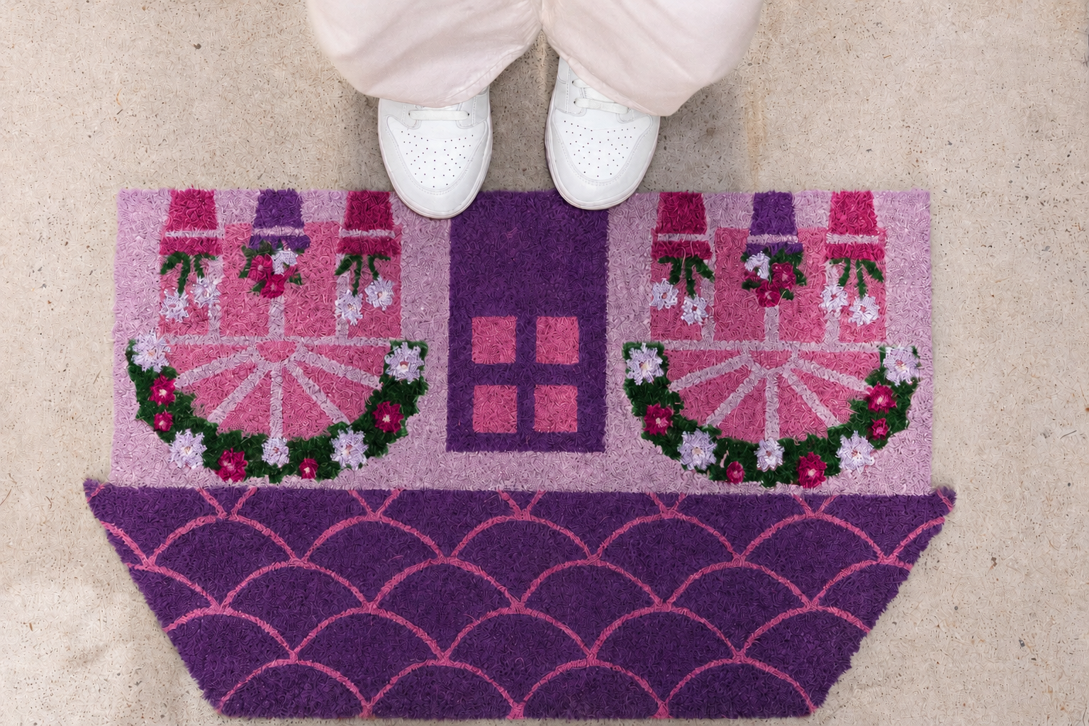 Top view of a person standing on a colorful, decorative rug with white sneakers, tan floor, and light pink pants.