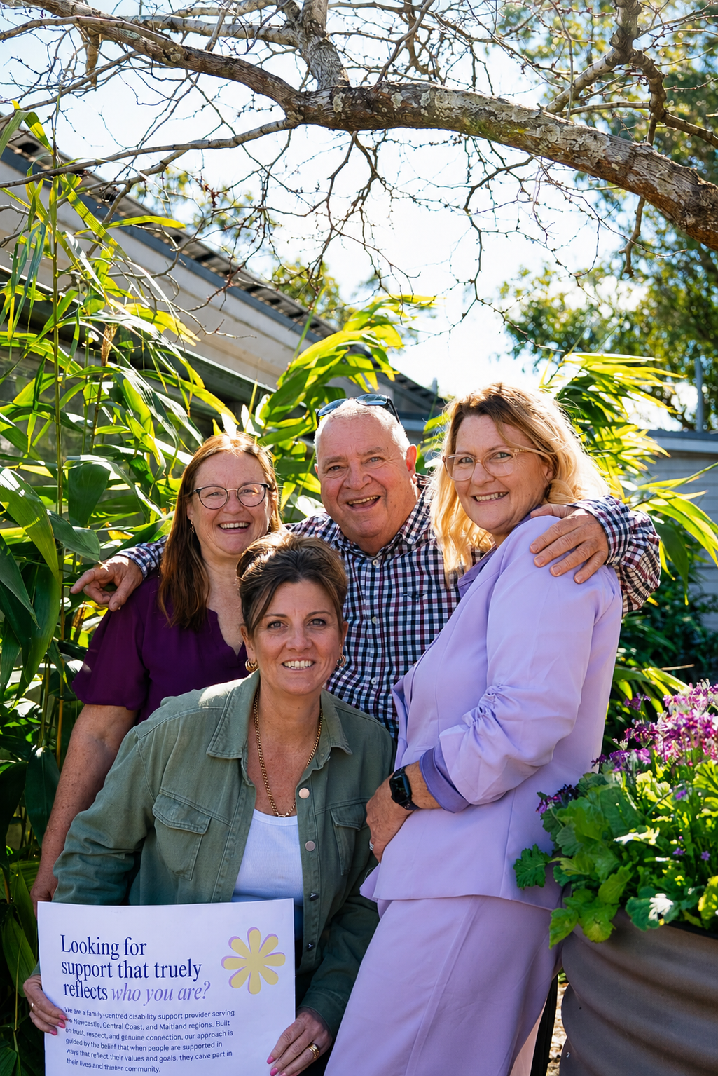 A group of four people, three women and one man, smiling and posing together outdoors with greenery and a blue sky in the background. One woman is holding a sign about support and disability services.