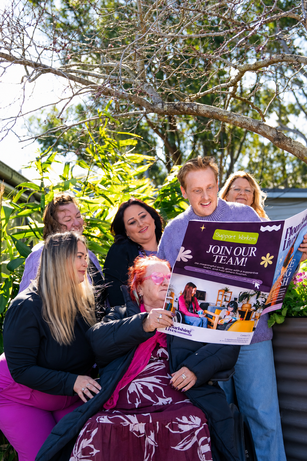 Group of women and a man gathered outdoors around an elderly woman in a wheelchair, looking at a colorful brochure, with trees and plants in the background.