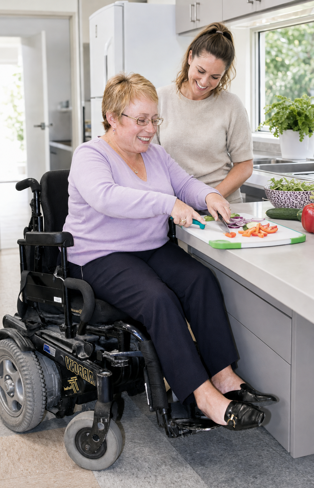 A woman in a wheelchair cooking vegetables with a caregiver in a bright kitchen.