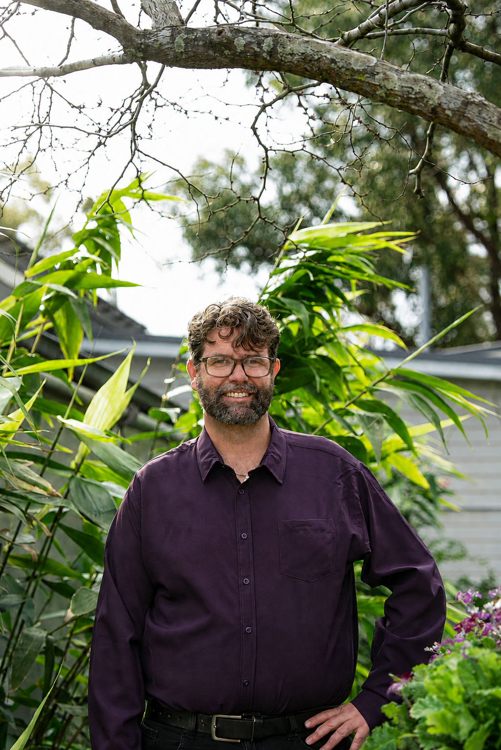 A smiling man with glasses and a beard standing outdoors among green plants and foliage, with a tree branch overhead and a house in the background.