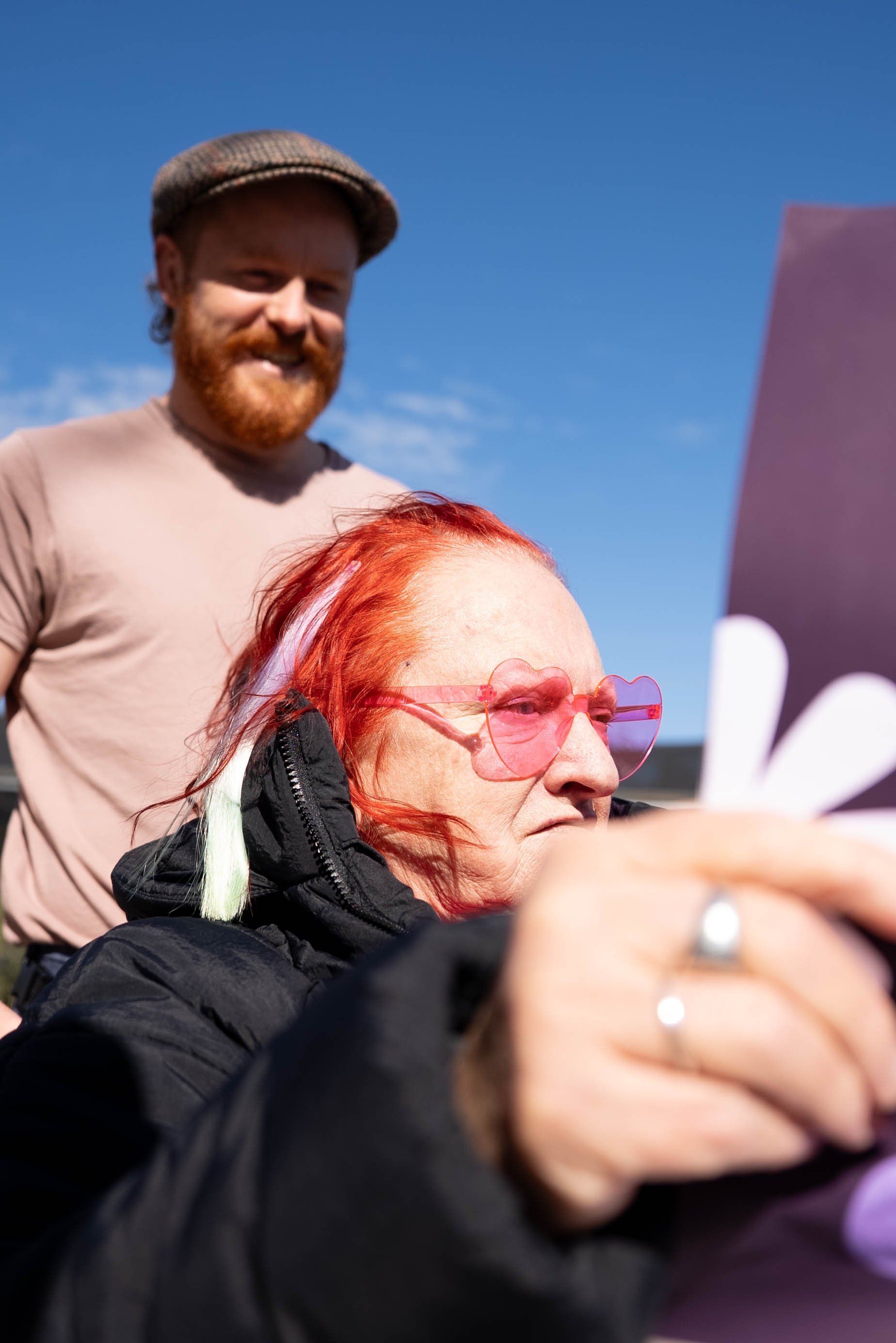A man with a red beard and a woman with red hair and pink sunglasses are outdoors with a blue sky. The man is smiling and wearing a brown cap and beige shirt; the woman is focused on a device she is holding.