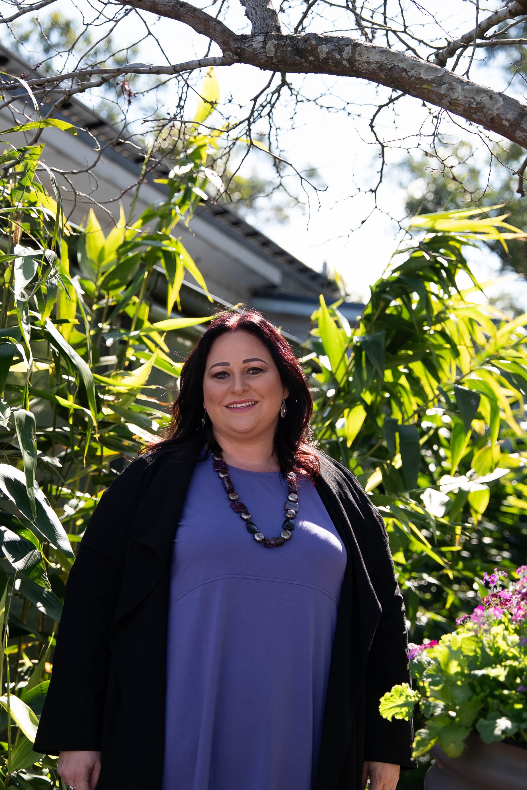 A woman with dark hair and a necklace standing outdoors among green plants, smiling at the camera, with a house and tree branches in the background.