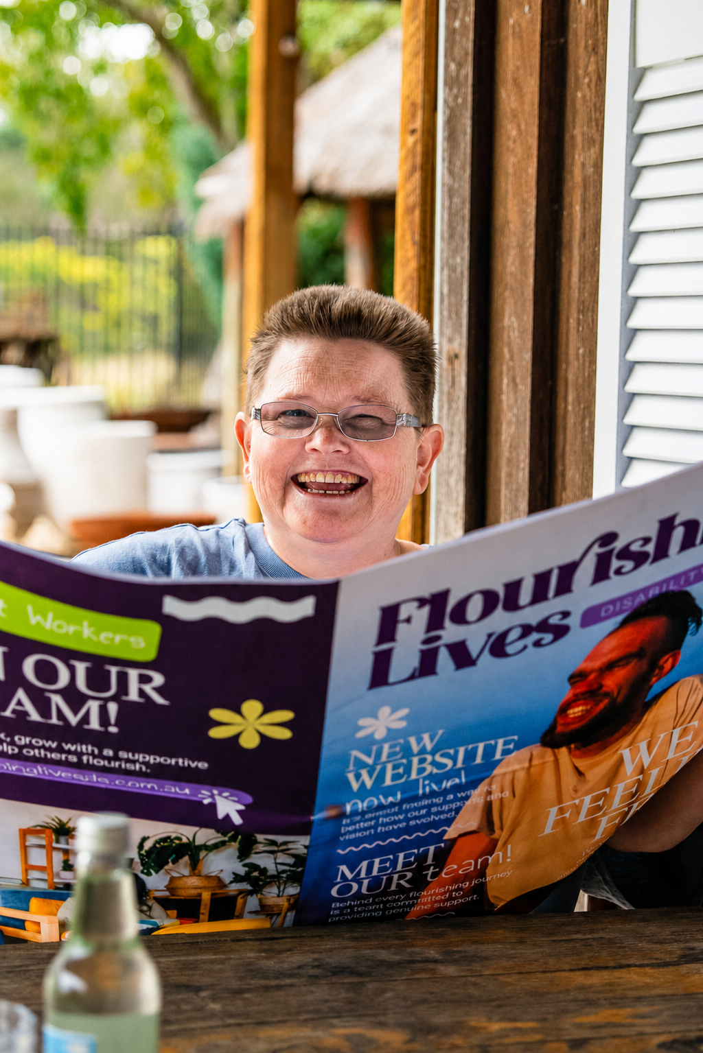 A joyful person with short hair and glasses smiles while reading a colorful magazine or flyer outside on a wooden table.