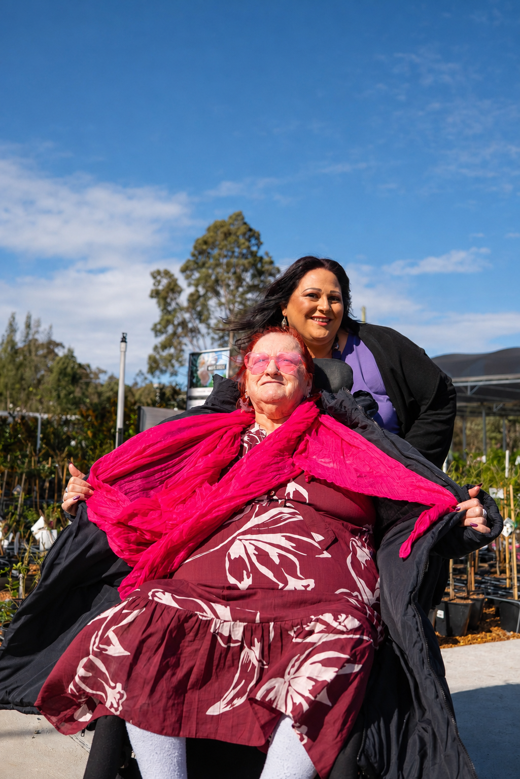 Two women, one elderly in a wheelchair with vibrant red hair, pink glasses, wearing a red and pink floral dress with a red scarf, and the other middle-aged with long dark hair, smiling, standing outside on a sunny day with blue sky and trees in the background.