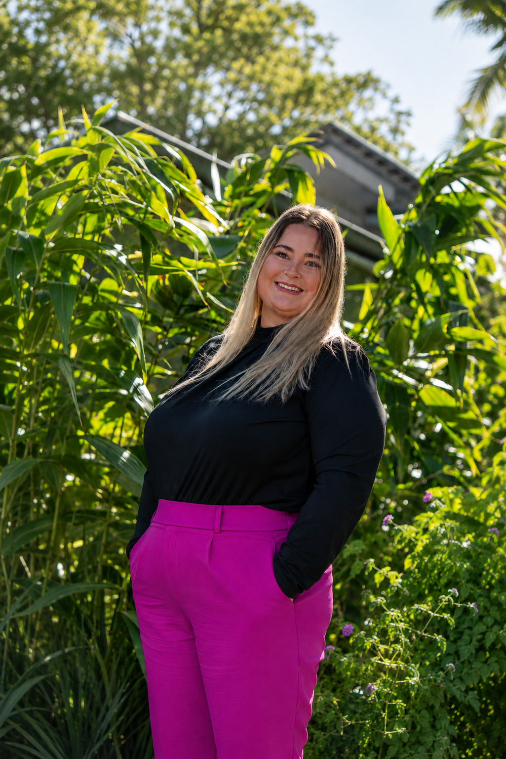 A woman with long blonde hair smiling outdoors next to tall green plants, wearing a black top and bright pink pants.