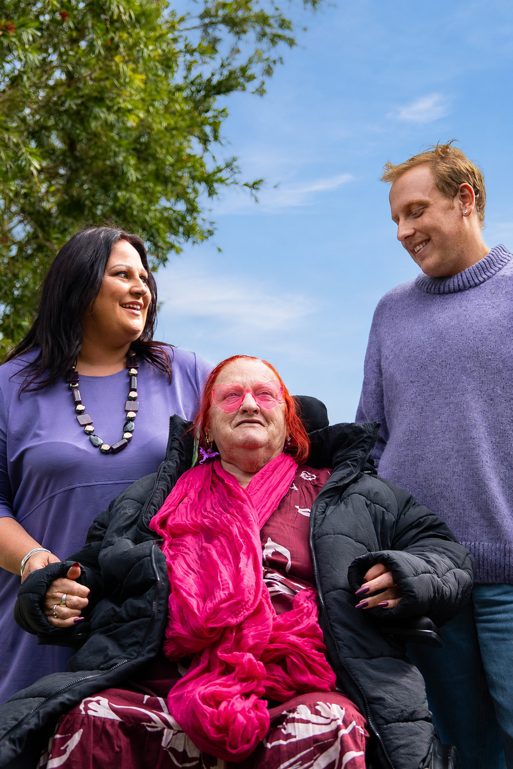 Three people outdoors, two standing and smiling at an elderly woman in a wheelchair, who is wearing pink glasses and a bright pink scarf, with a tree and blue sky in the background.