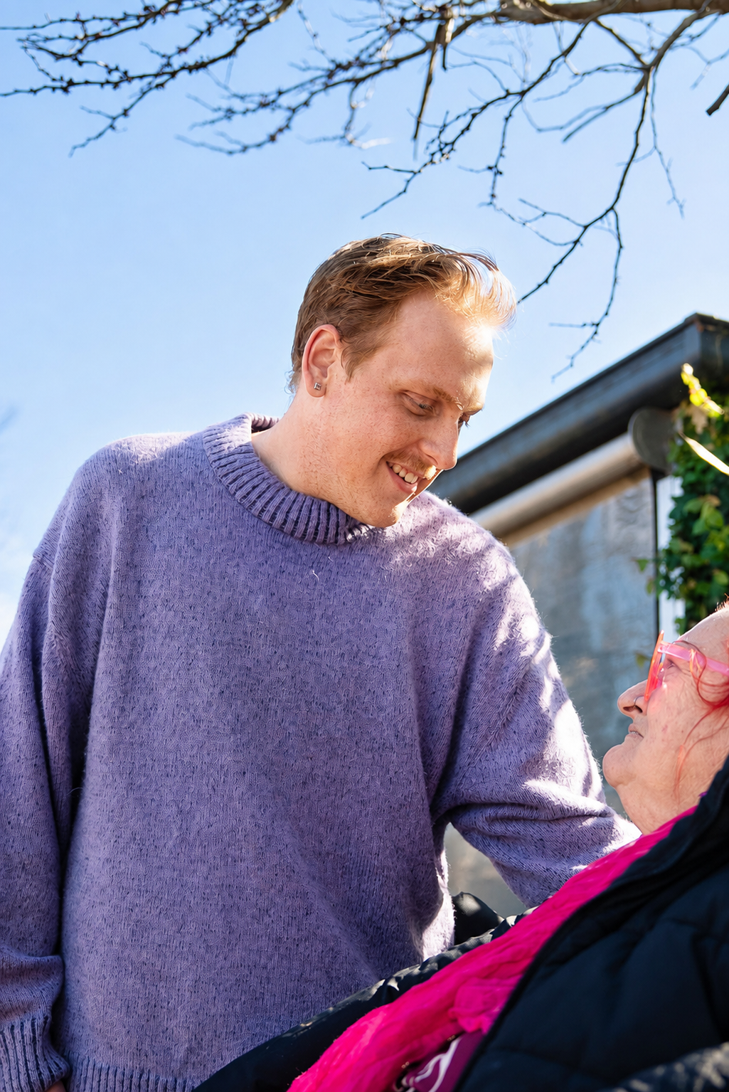 A young man with light skin, blonde hair, and wearing a purple sweater is smiling and looking down at an elderly woman with glasses and red hair, who is sitting in a wheelchair outdoors on a sunny day with a blue sky and leafless tree branches overhead.