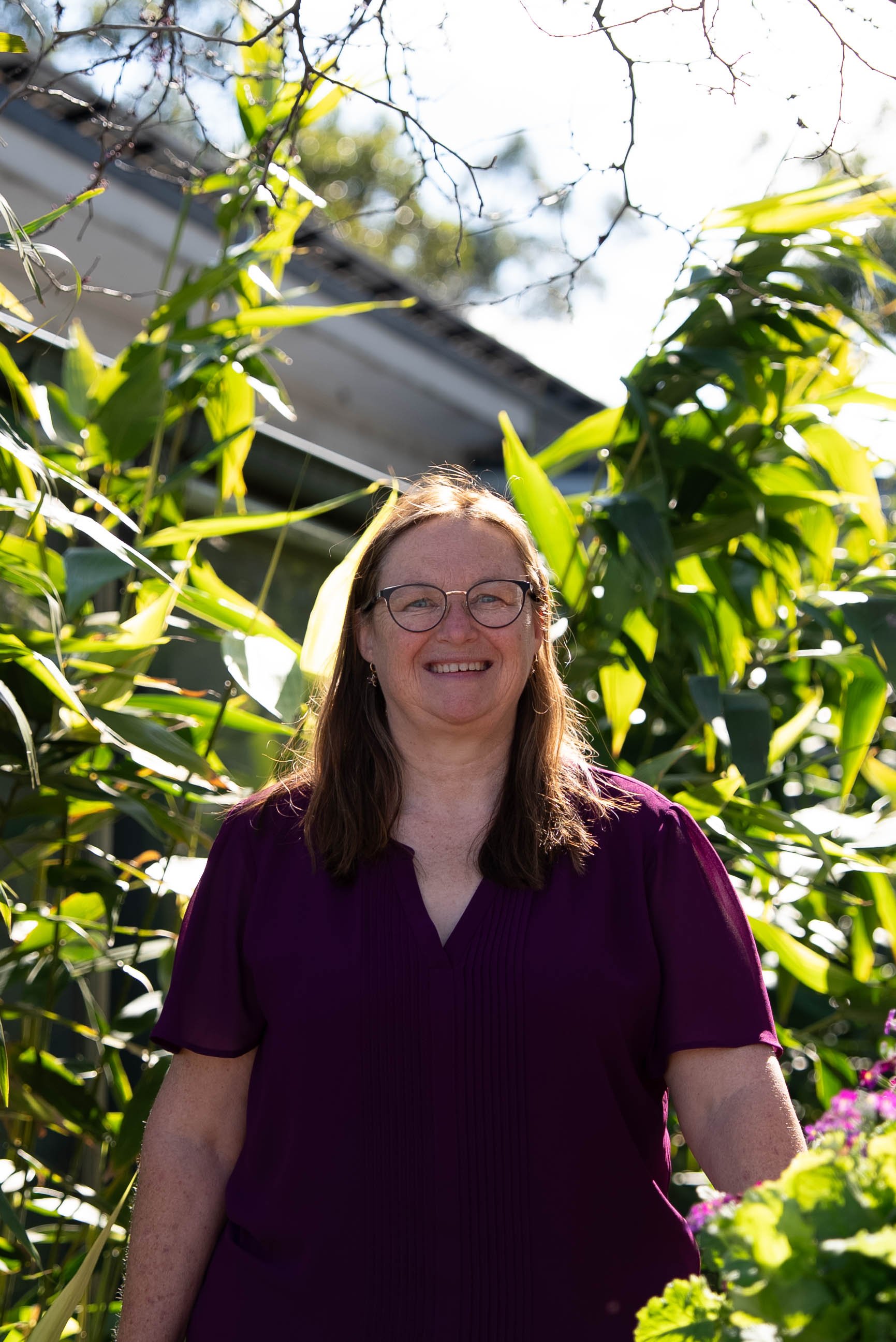 A woman with glasses and long brown hair smiling outdoors among tall green plants on a sunny day.