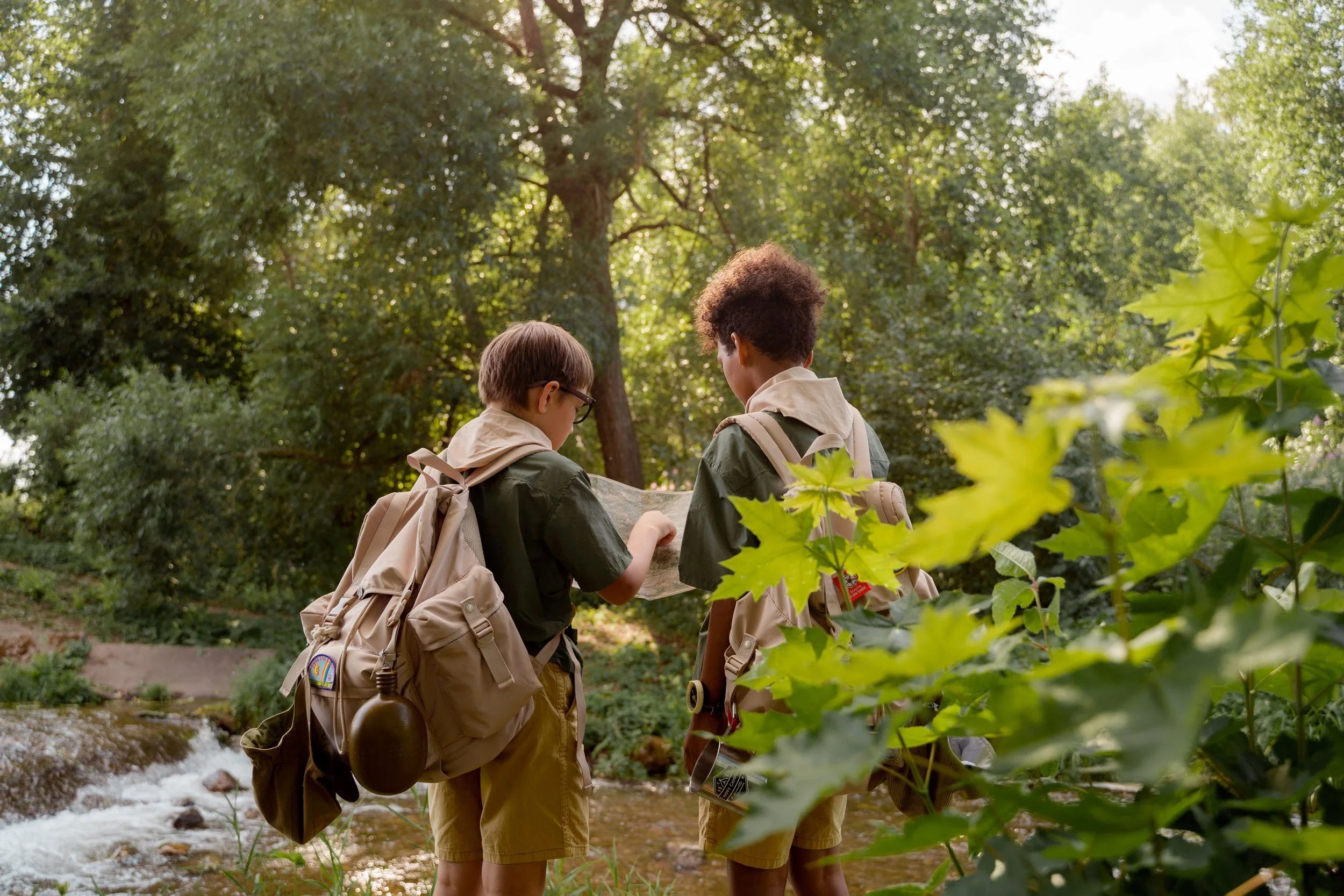 Two campers with backpacks hike along a wooded stream while checking directions, reflecting summer camp scholarships that help more children access enriching outdoor experiences.