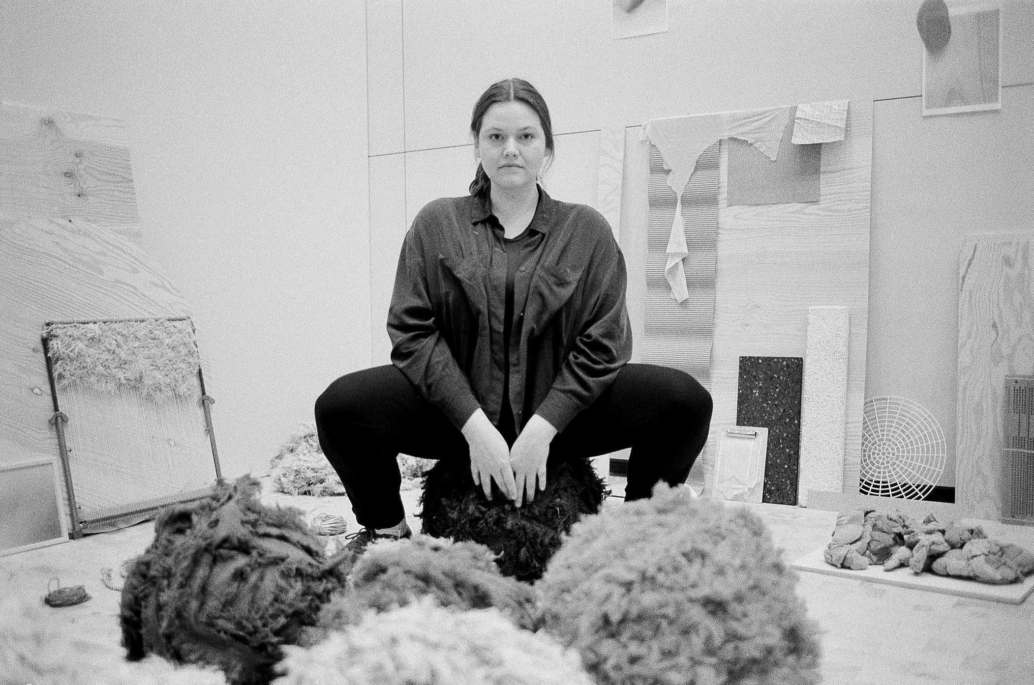 Bilde av Katrin Rusten som sitter på en krakk, sort hvitt bilde, rommet er fyllt med kunst