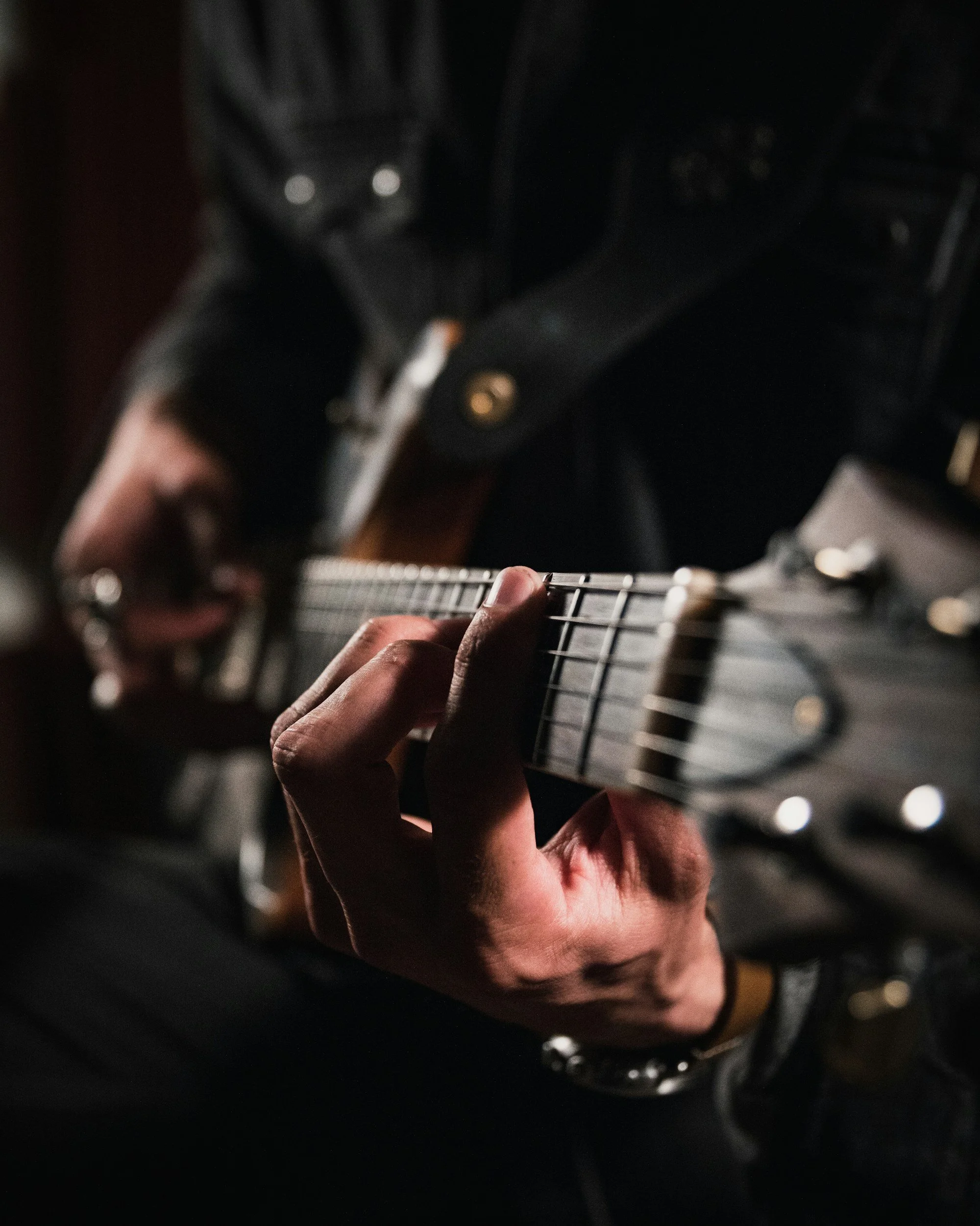 Close-up of a person's hand pressing the strings on an electric guitar's fretboard, with a blurred background of a drum set.