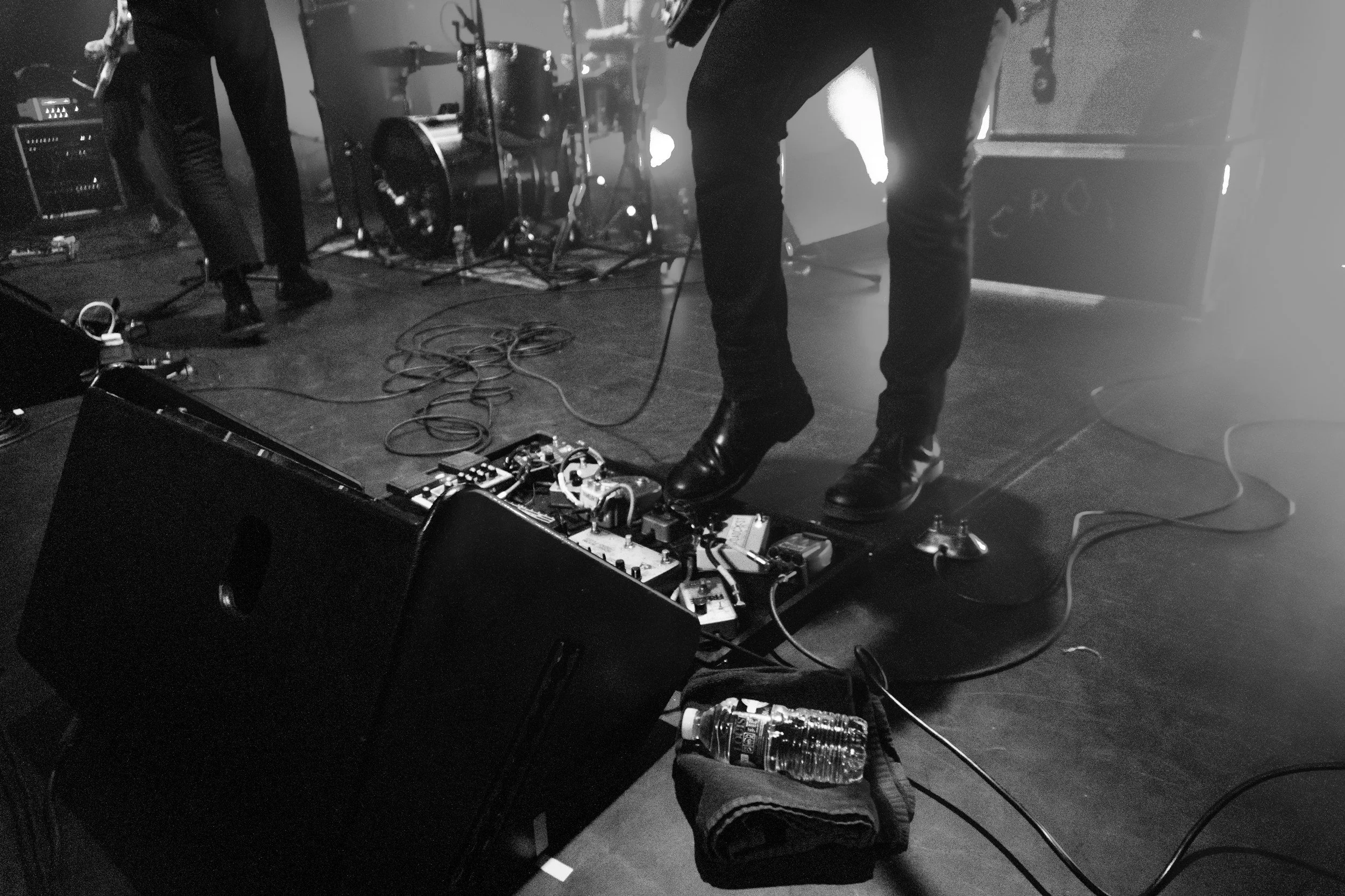 Black and white photo of a band on stage, showing the lower half of musicians, various equipment, and tangled cables.