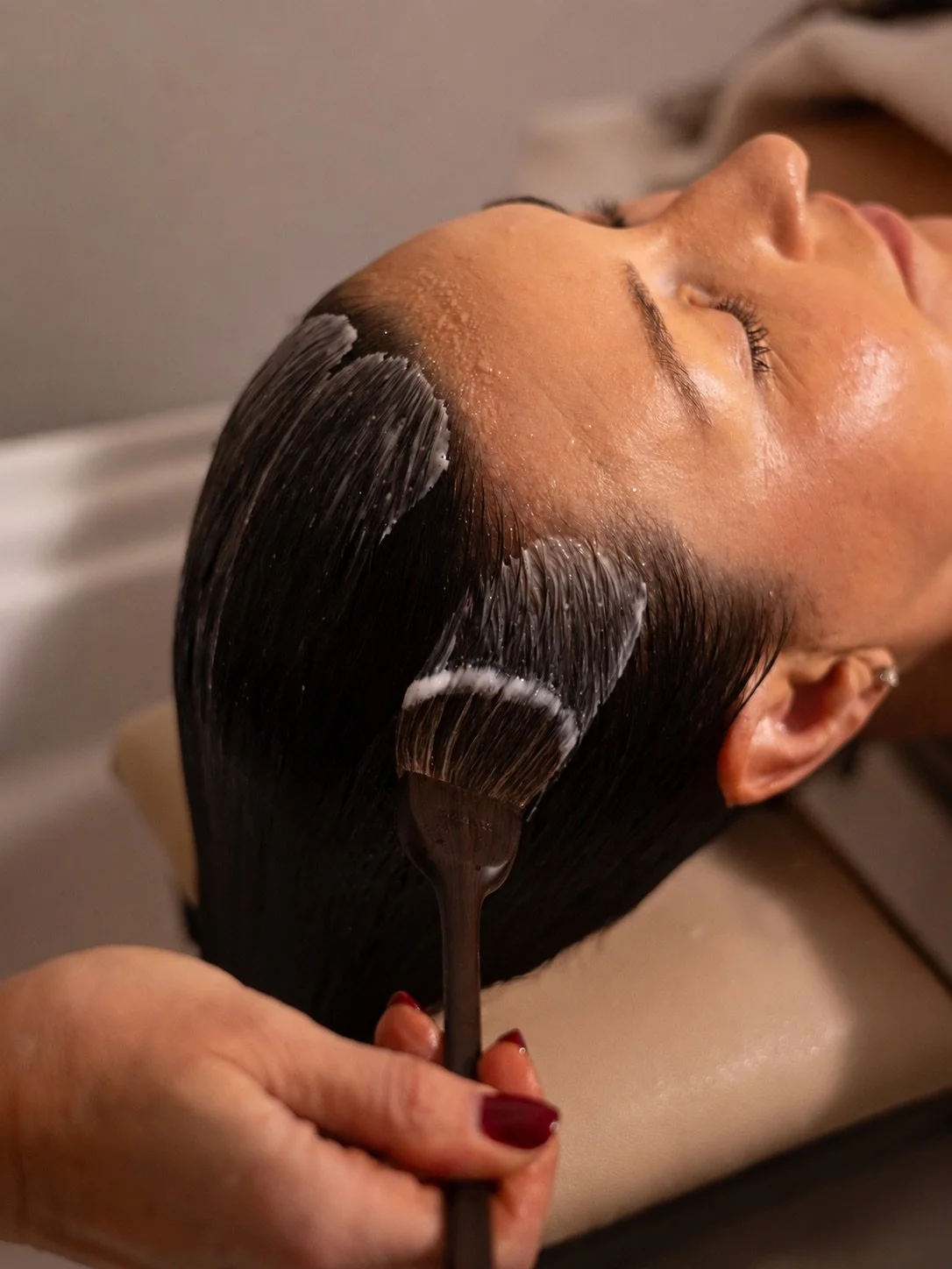 A woman receiving a hair treatment or scalp massage at a spa, with a stylist applying a dark liquid substance to her hair and scalp.