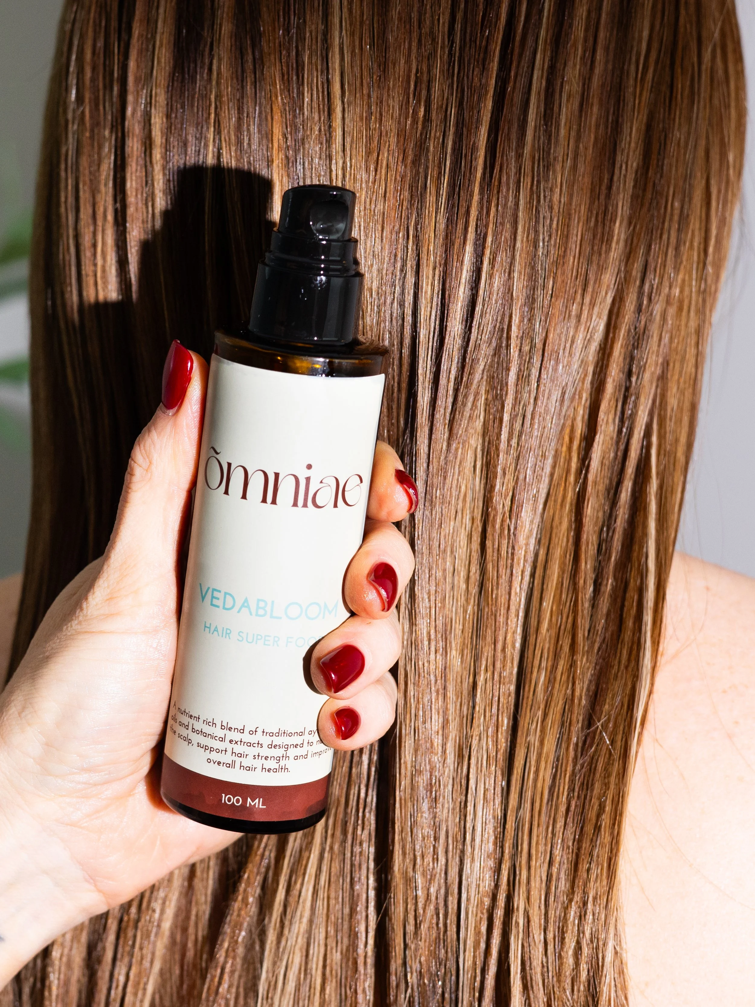 A hand with red-painted nails holding a brown bottle of hair care product labeled 'Omniage Vedabloom Hair Super Food,' positioned against a background of straight, shiny brown hair.