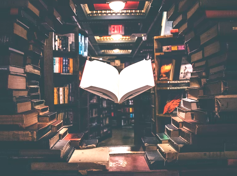 A floating open book illuminated in a bookstore or library surrounded by shelves of stacked books.