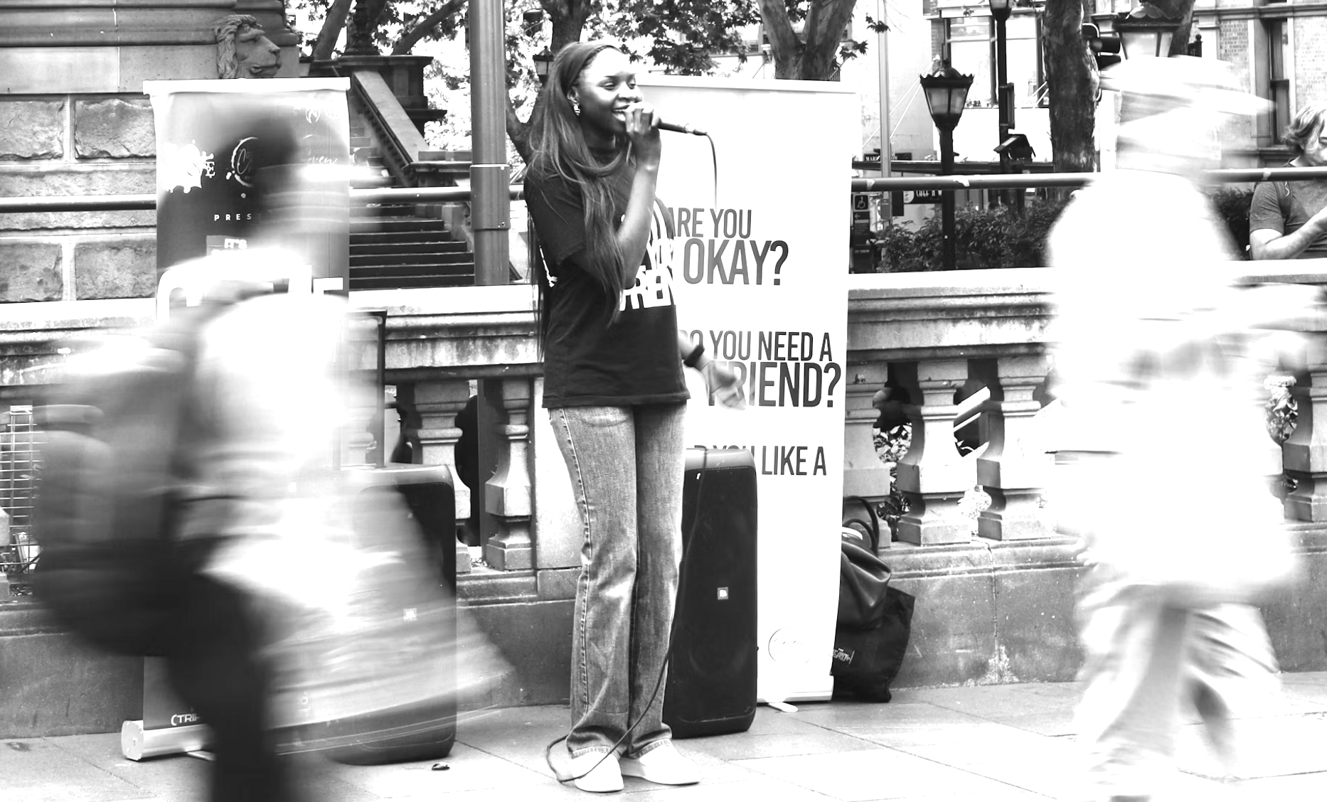 A woman speaking at an outdoor event, holding a microphone, with a banner behind her. The scene is on a city street with blurred pedestrians passing by.
