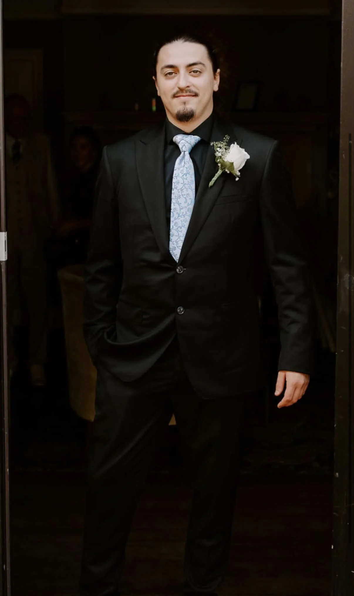 A man dressed in a black suit with a white boutonniere, gray patterned tie, and black dress shirt standing in an elevator