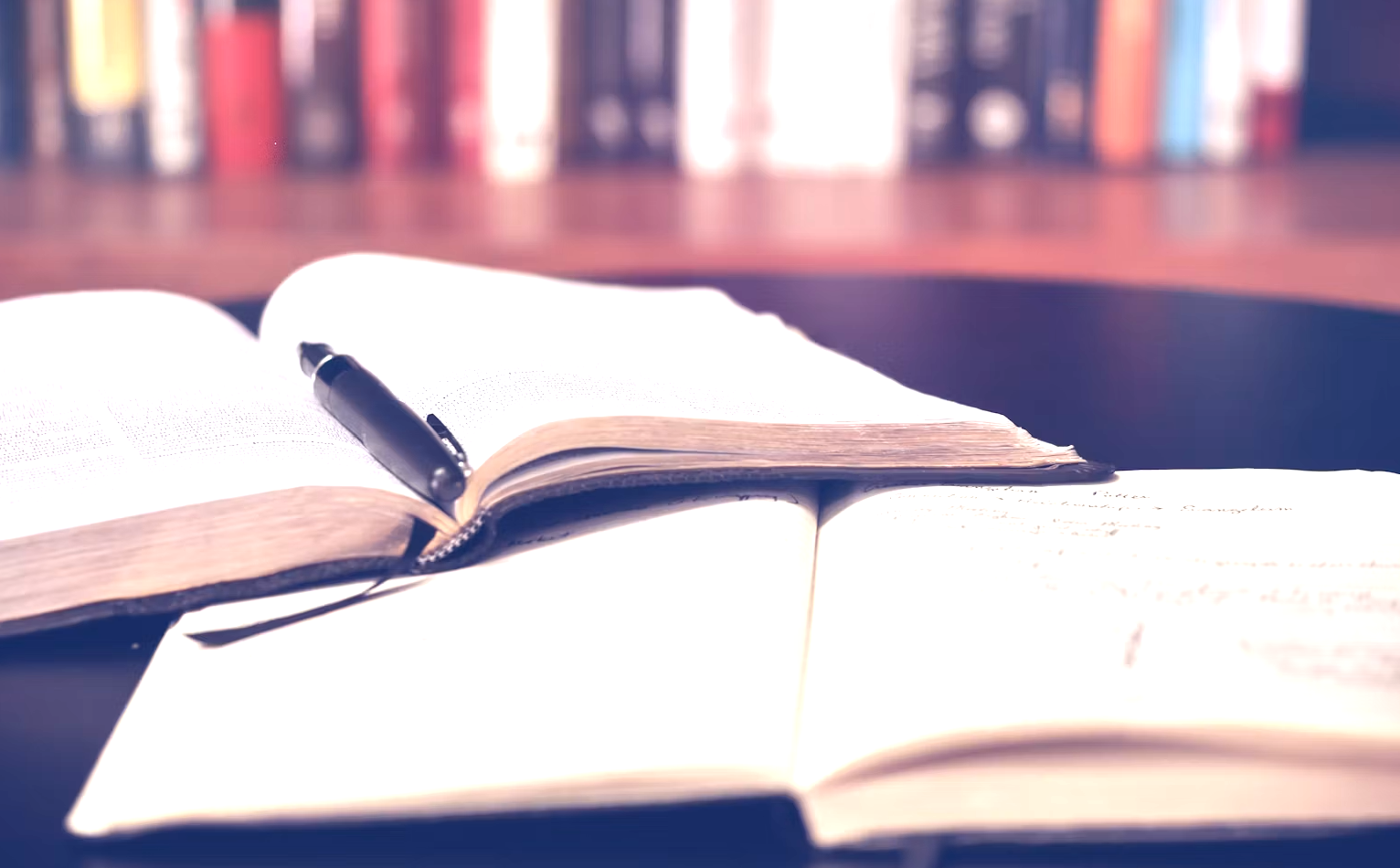 Open book with a pen on top, placed on a table with a background of blurry bookshelves.