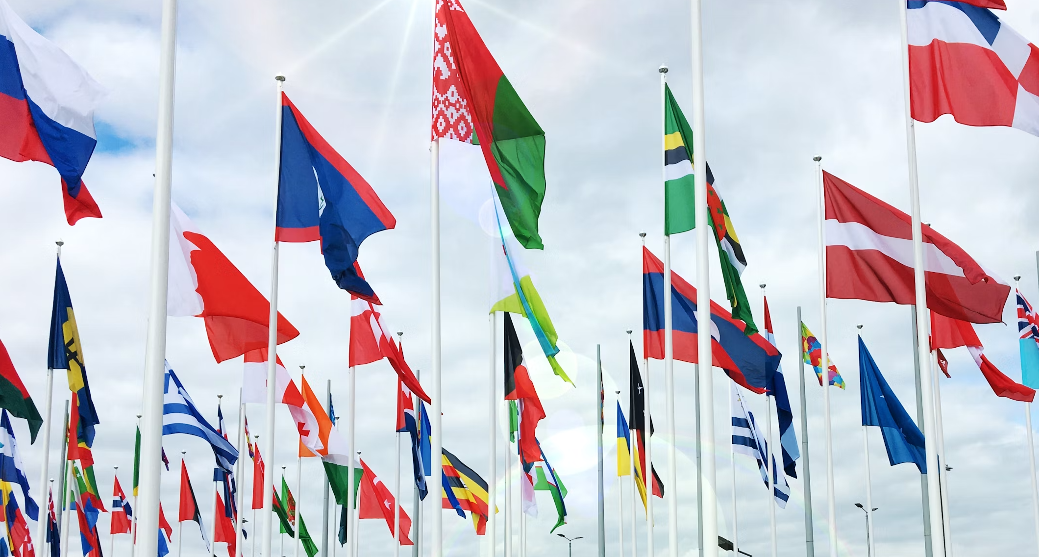 A large collection of international flags on flagpoles flying in the wind against a cloudy sky.