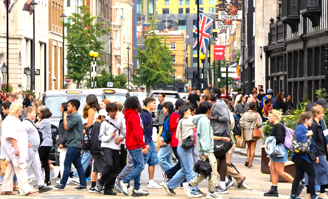Crowd of people crossing a city street, with buildings, flags, and trees in the background.