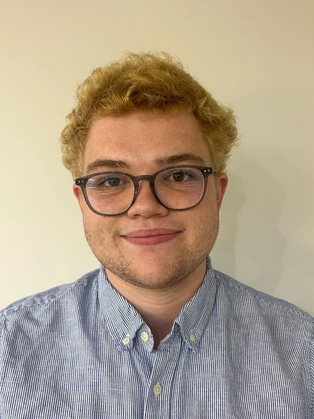 A young man with curly blonde hair, wearing round glasses and a light blue striped button-up shirt, smiling in front of a plain light-colored wall.