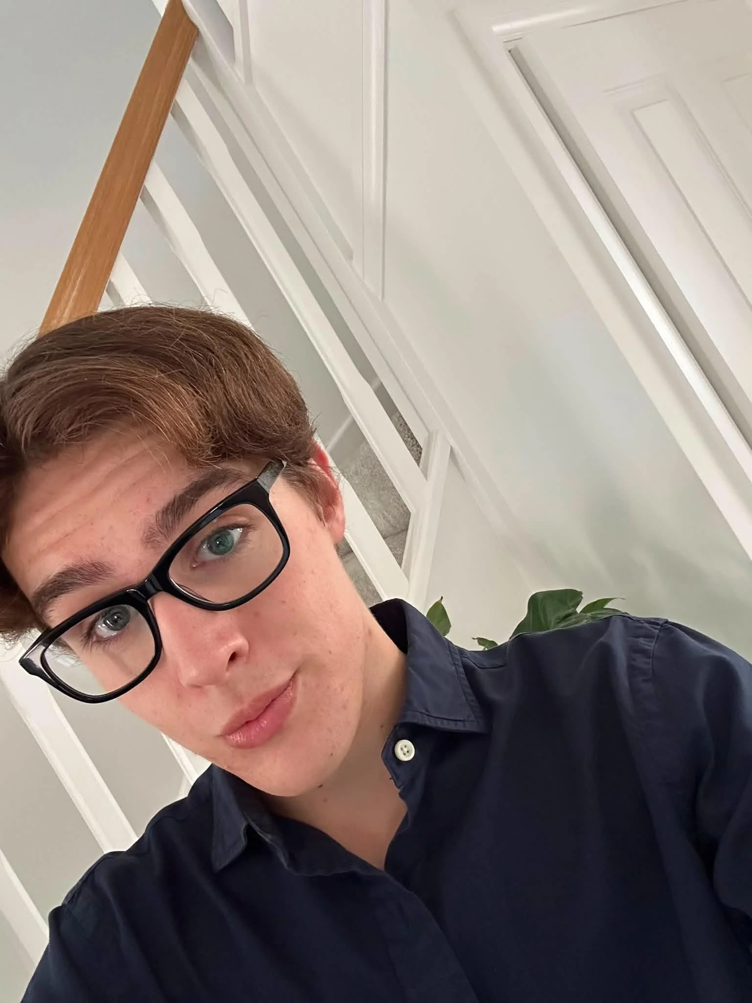 Close-up of a young man with red hair and glasses, wearing a navy blue shirt, indoors near white walls and cabinets, with a green plant in the background.