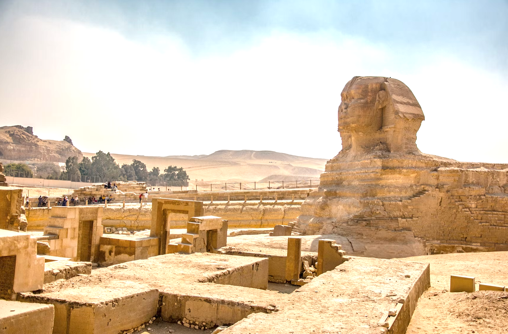 The Great Sphinx of Giza in Egypt with desert landscape and pyramid ruins in the background, several tourists exploring the site.